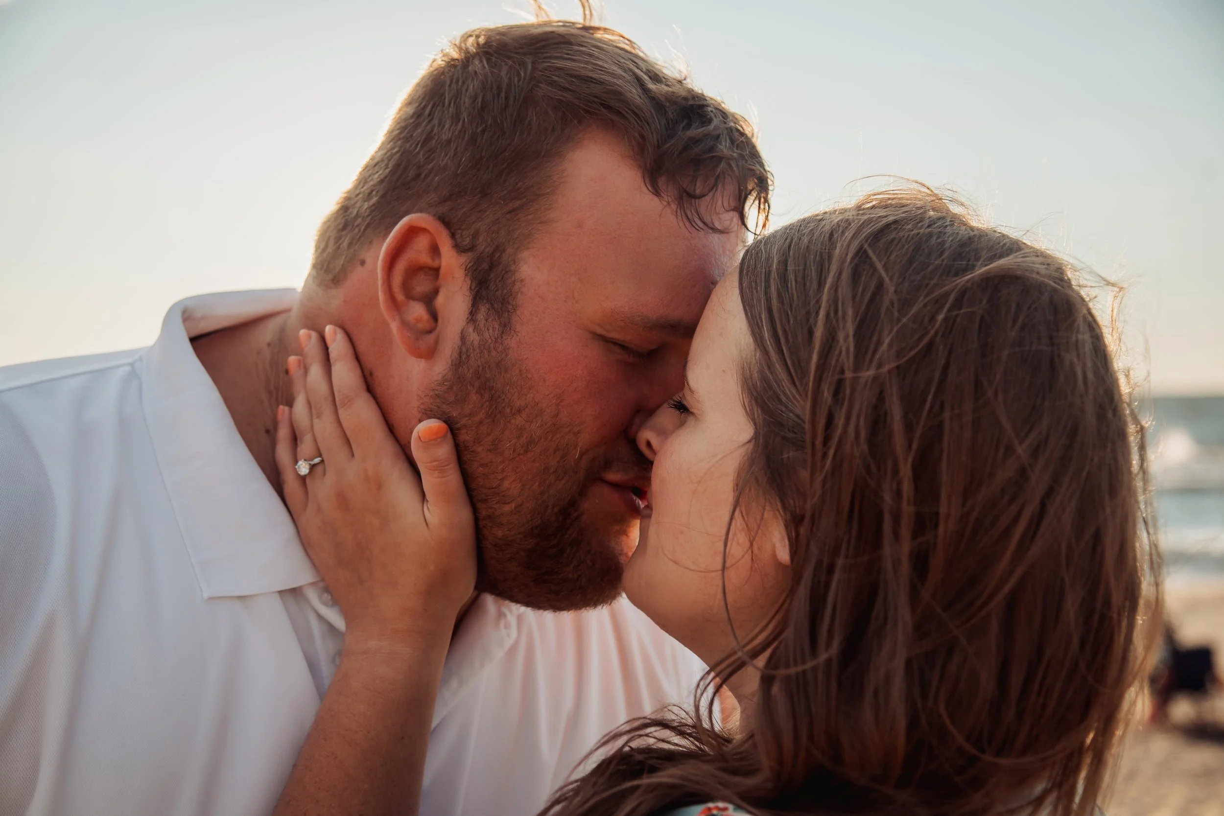 A couple sharing a romantic kiss at the beach with an ocean background during sunset.