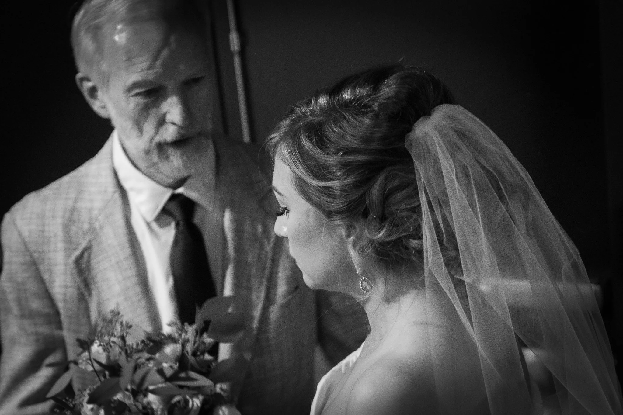 A bride with a veil and earrings looking down, standing beside an older man in a suit, holding a bouquet, in a black-and-white photograph.