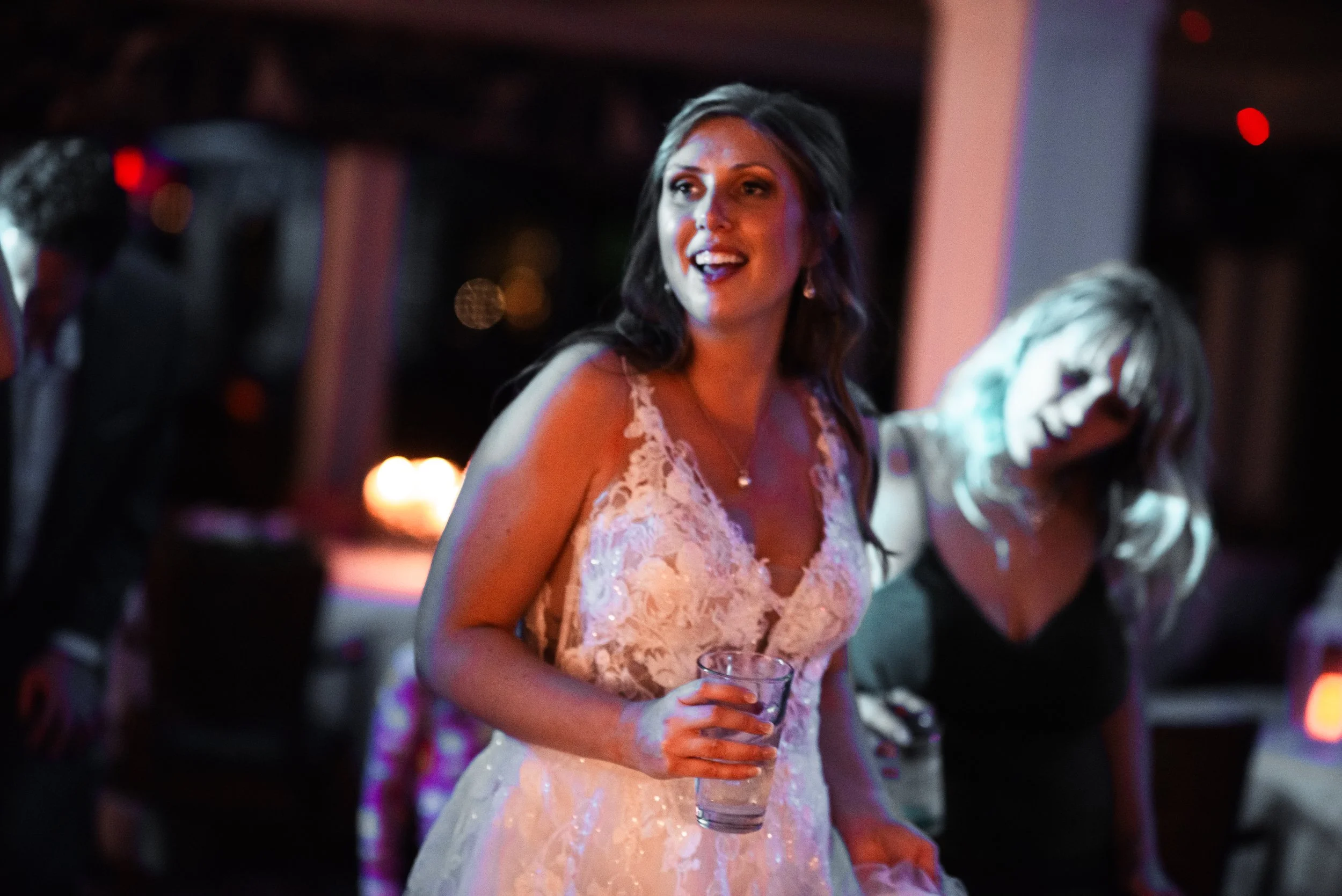 A woman in a white wedding dress holding a glass of water at a wedding reception, smiling and talking, with other guests in the background.