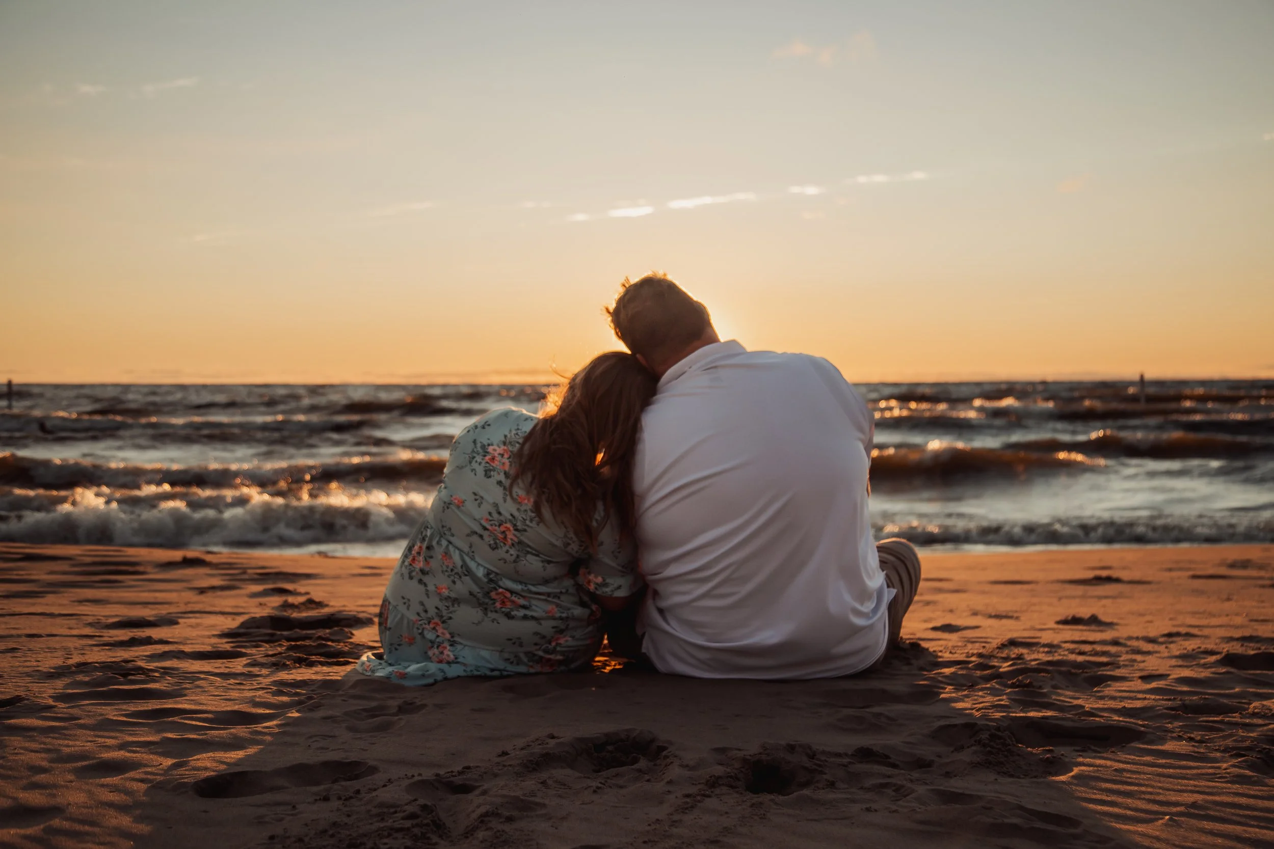 A couple sitting on the beach during sunset, with the woman leaning her head on the man's shoulder.