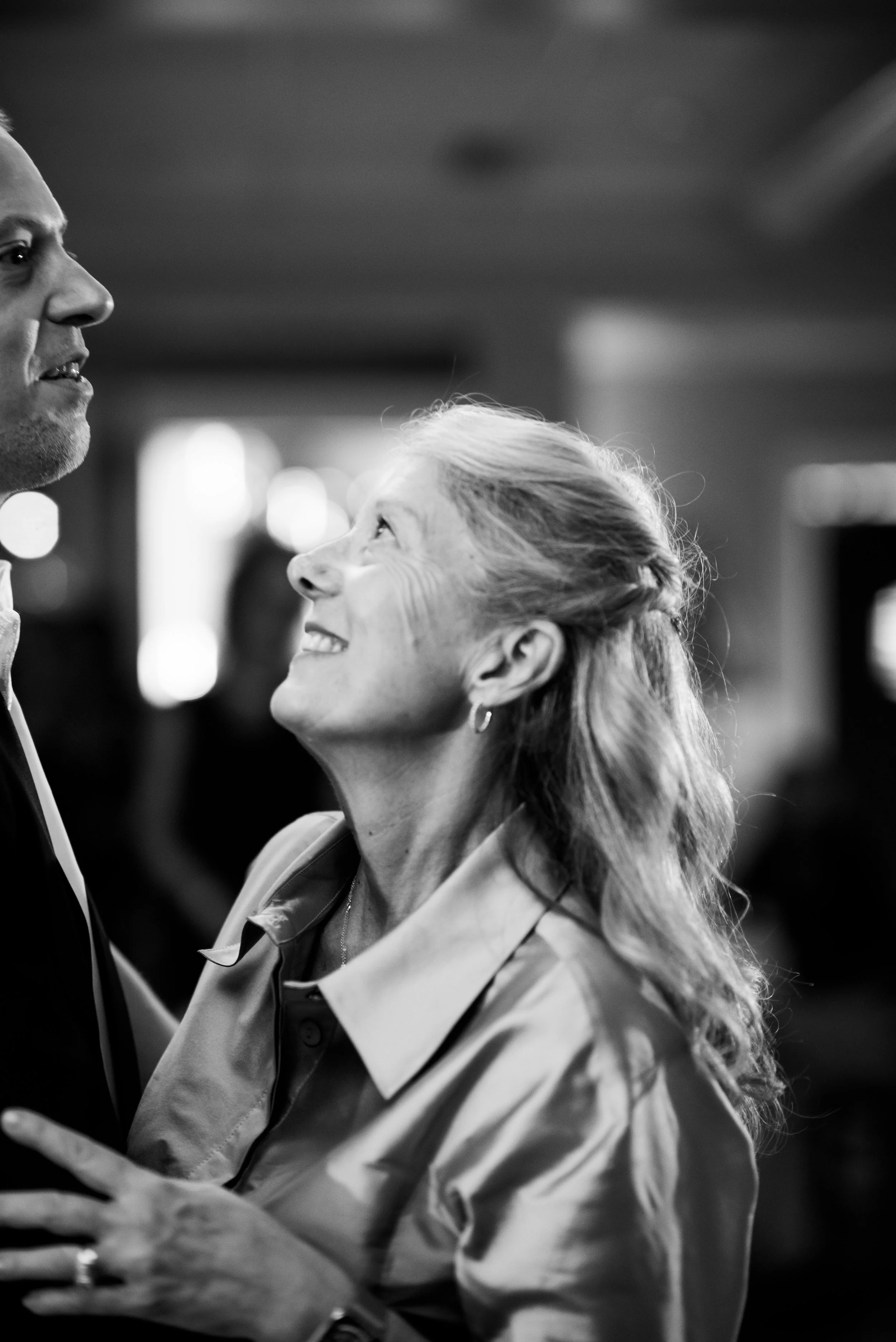 A black and white photo of a woman smiling up at a man, both in business attire, with a warm and intimate expression, indoors with blurred background lights.