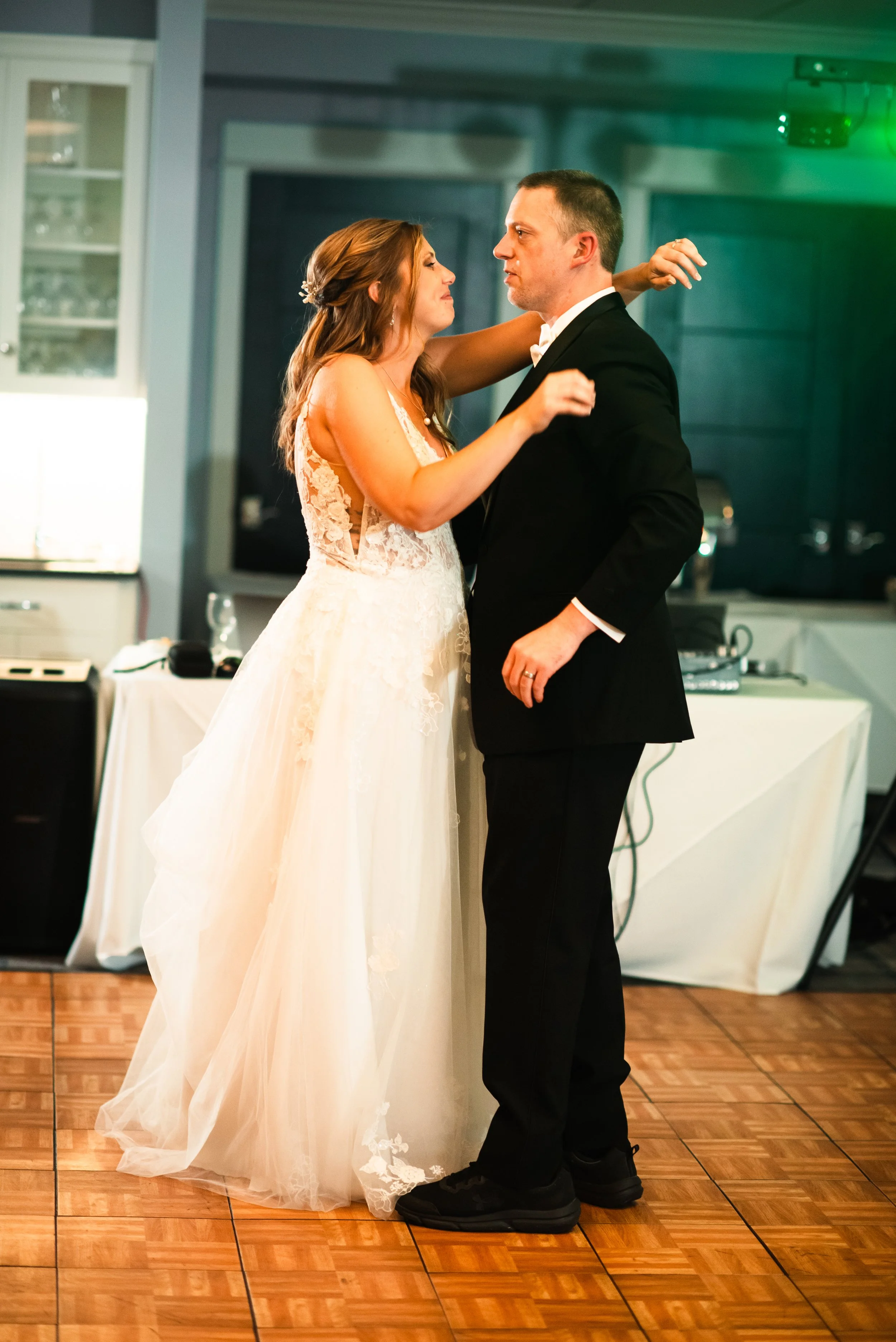 Bride and groom dancing closely at their wedding reception, with the bride in a white lace gown and the groom in a black tuxedo, on a wooden dance floor.