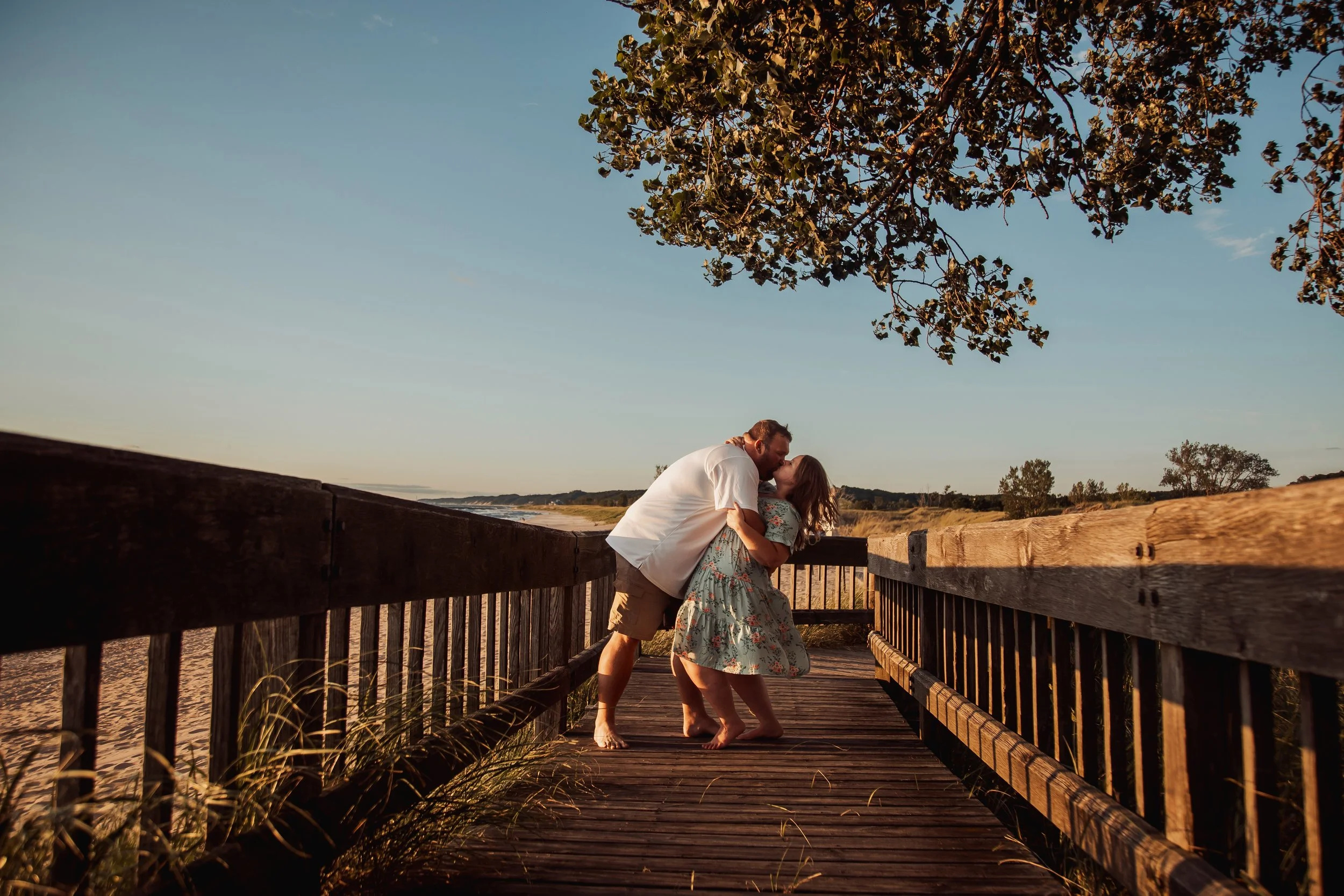 A couple kissing on a wooden boardwalk near the beach during sunset.