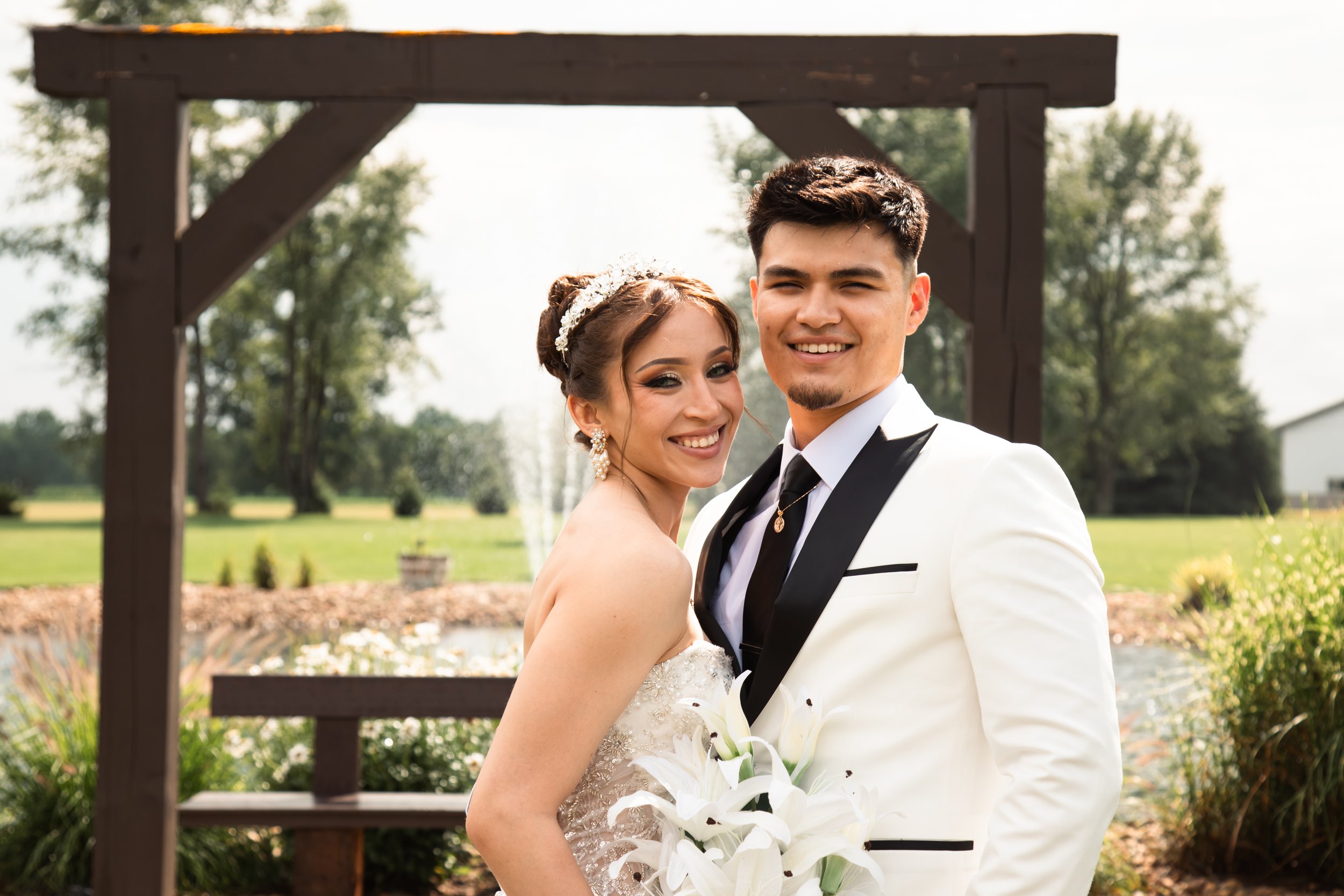 A newlywed couple stands outdoors, smiling at the camera. The bride wears a strapless wedding gown with lace details and holds a bouquet of white lilies. The groom wears a white tuxedo jacket with black lapels and a black tie, with a necklace visible