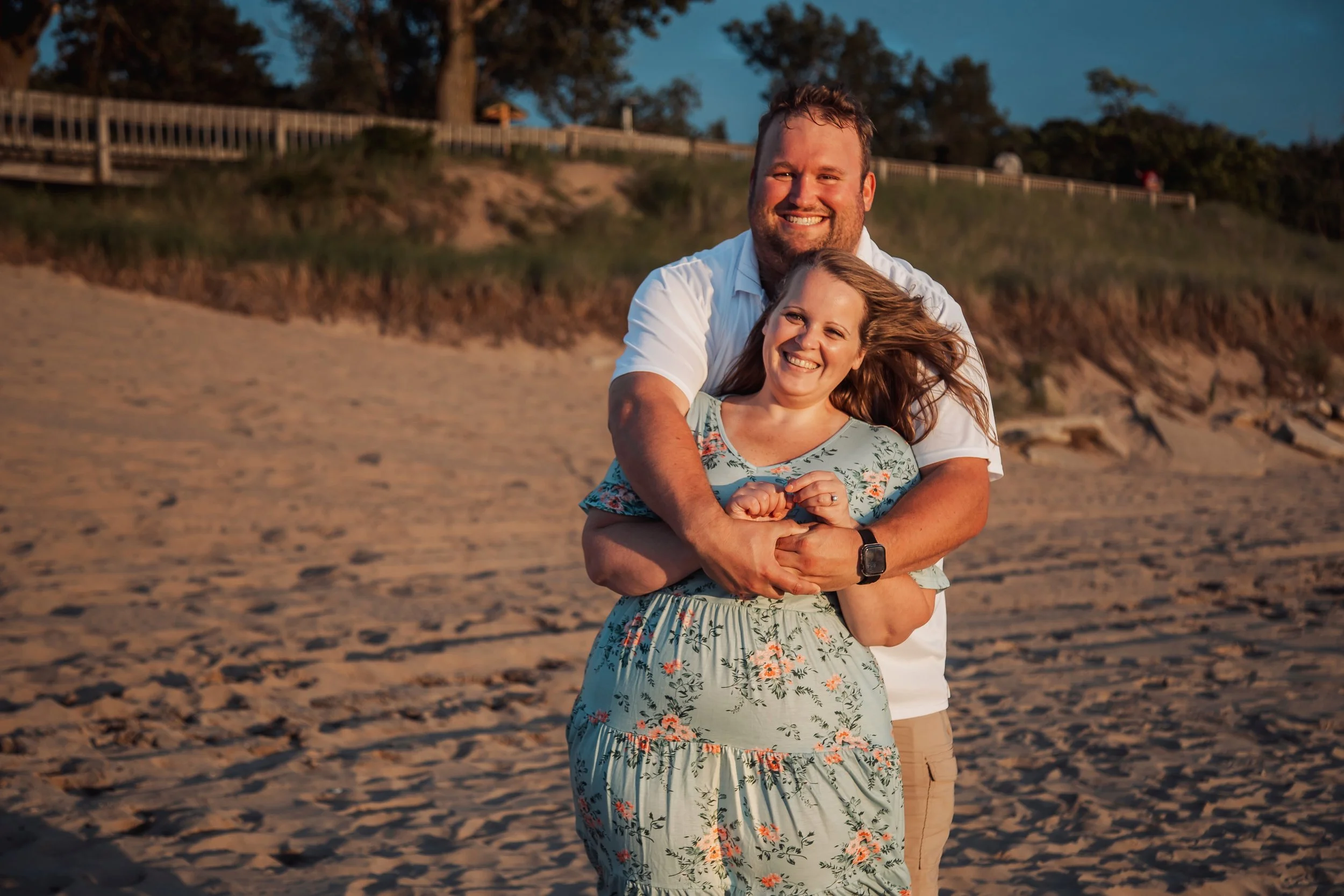 A smiling couple embracing on a sandy beach during sunset, with a wooden fence and grassy hill in the background.