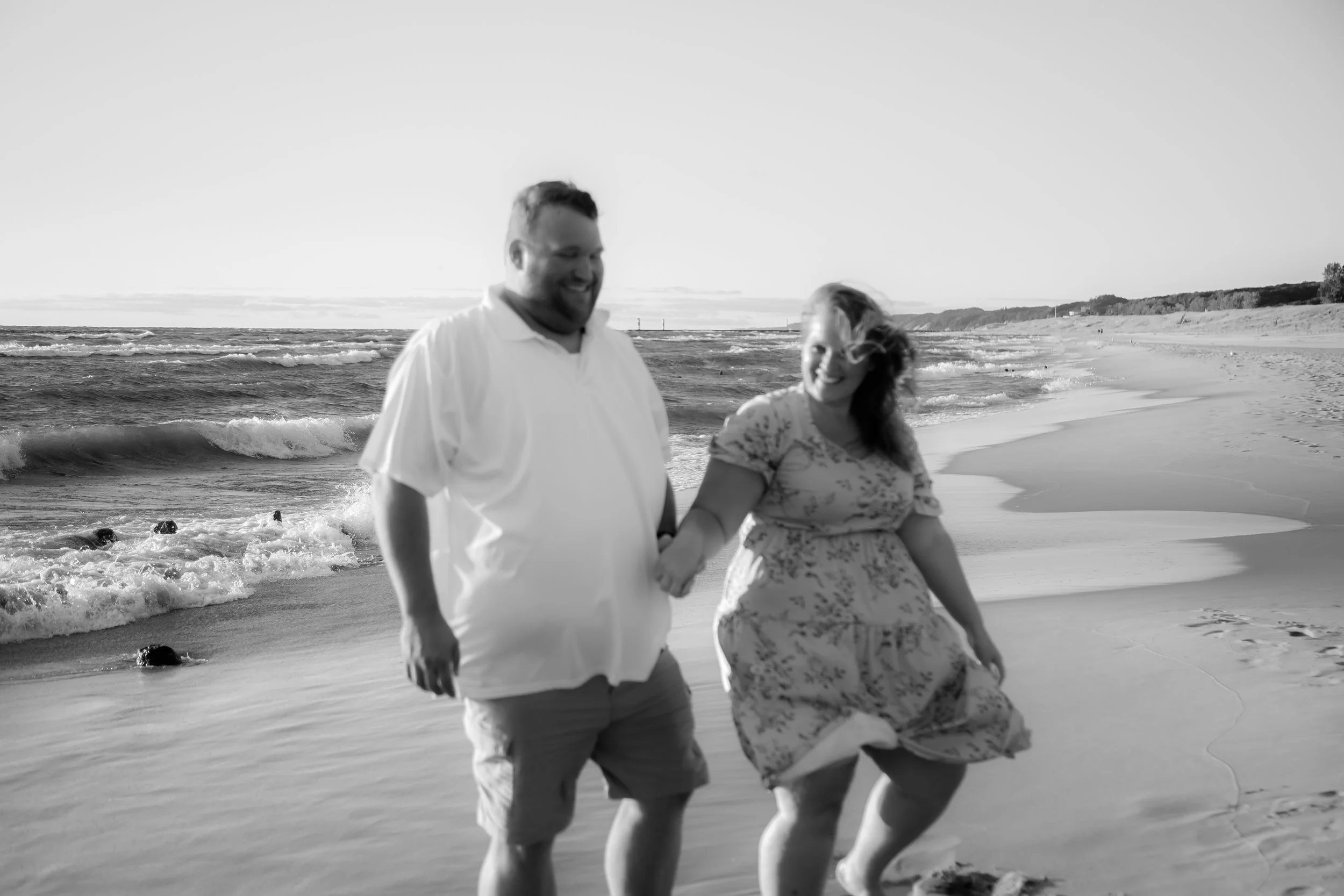 A couple walking hand-in-hand on a beach, smiling, with ocean waves and distant shoreline in the background, in black and white.