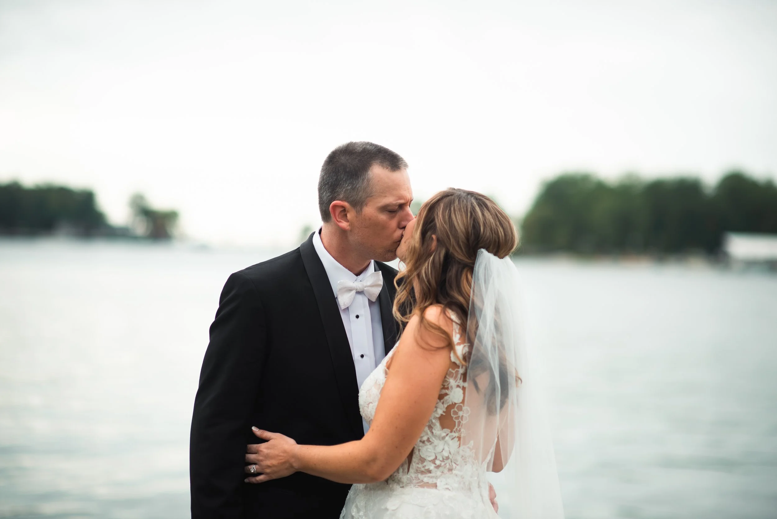 A newlywed couple sharing a kiss near a body of water, with trees and overcast sky in the background.