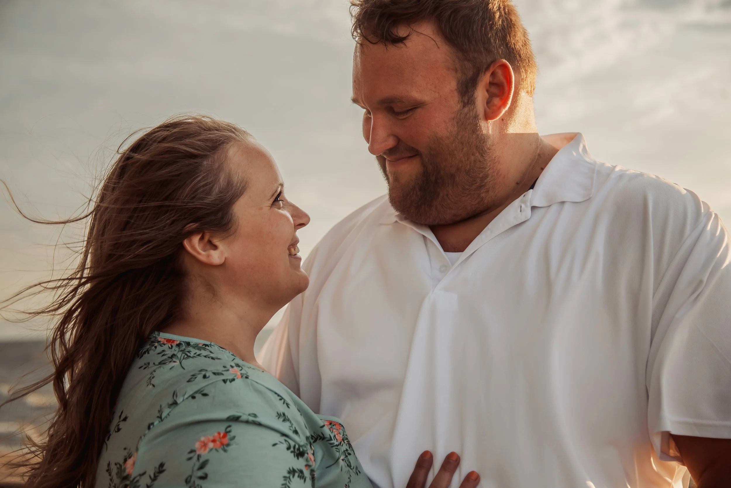 A couple looking at each other and smiling, outdoors during sunset.