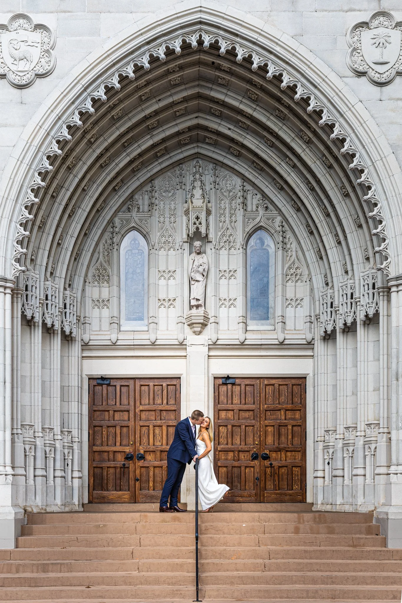 A couple in wedding attire standing on steps in front of a large church entrance with wooden doors and ornate stone architecture, kissing.