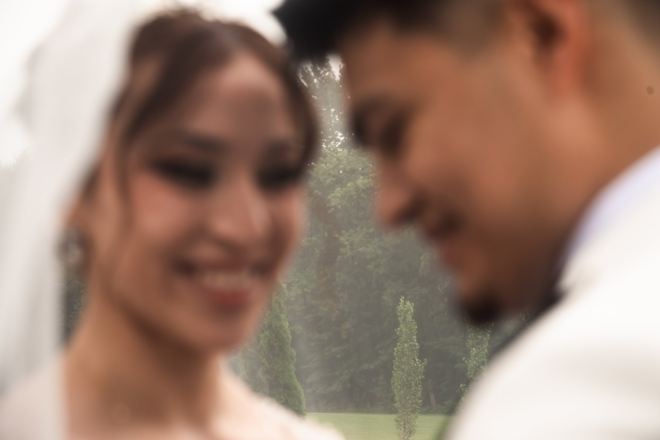 Blurred close-up of a smiling woman with long hair and a man with short hair, wearing formal attire, outdoors with trees in the background.