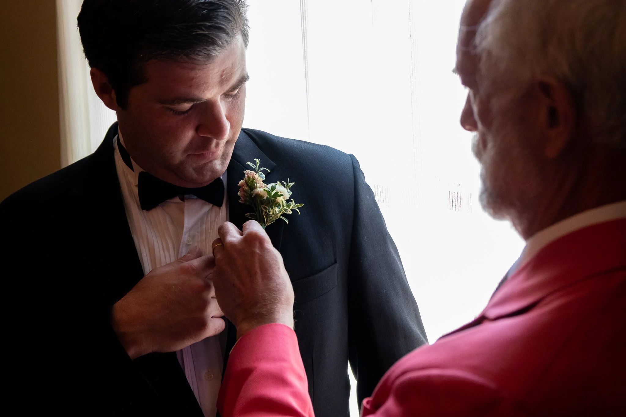 An elderly man pinning a boutonniere onto a young man in a tuxedo, likely during a wedding preparation.