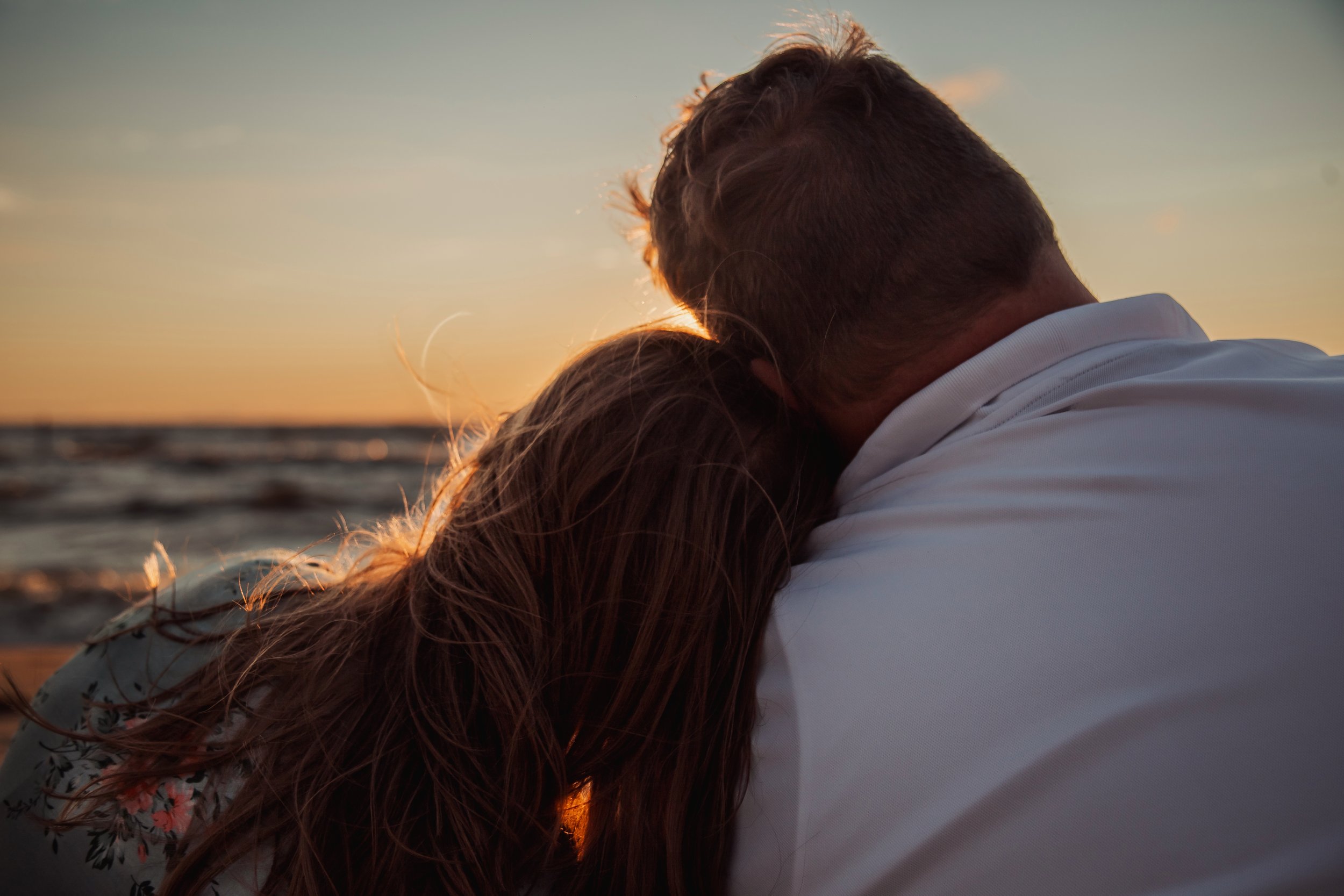 A couple embracing on the beach during sunset, with the woman's head resting on the man's shoulder, the ocean and sunset in the background.