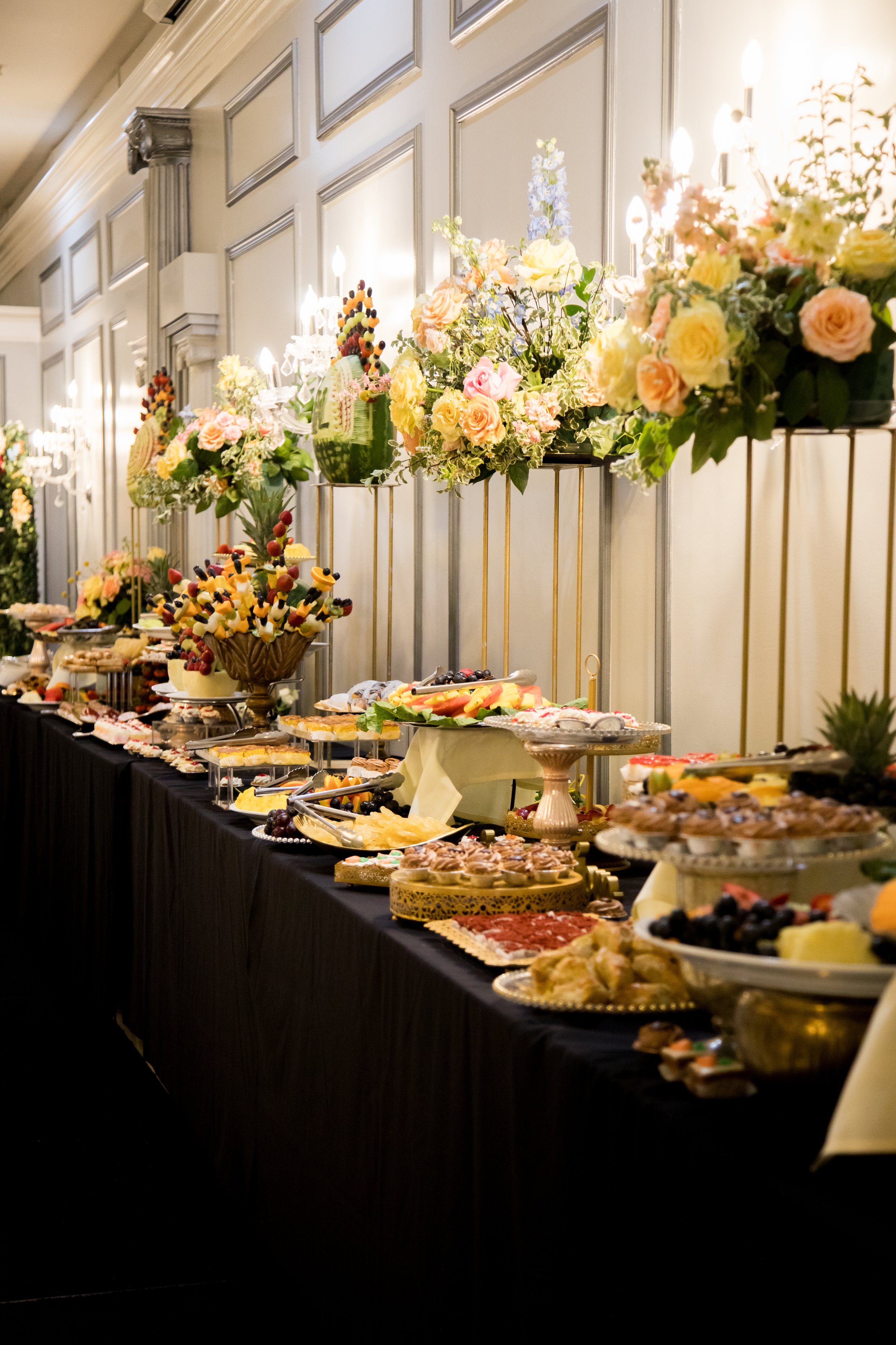 A decorated buffet table with an assortment of desserts and fruit, topped with floral arrangements in a banquet hall.