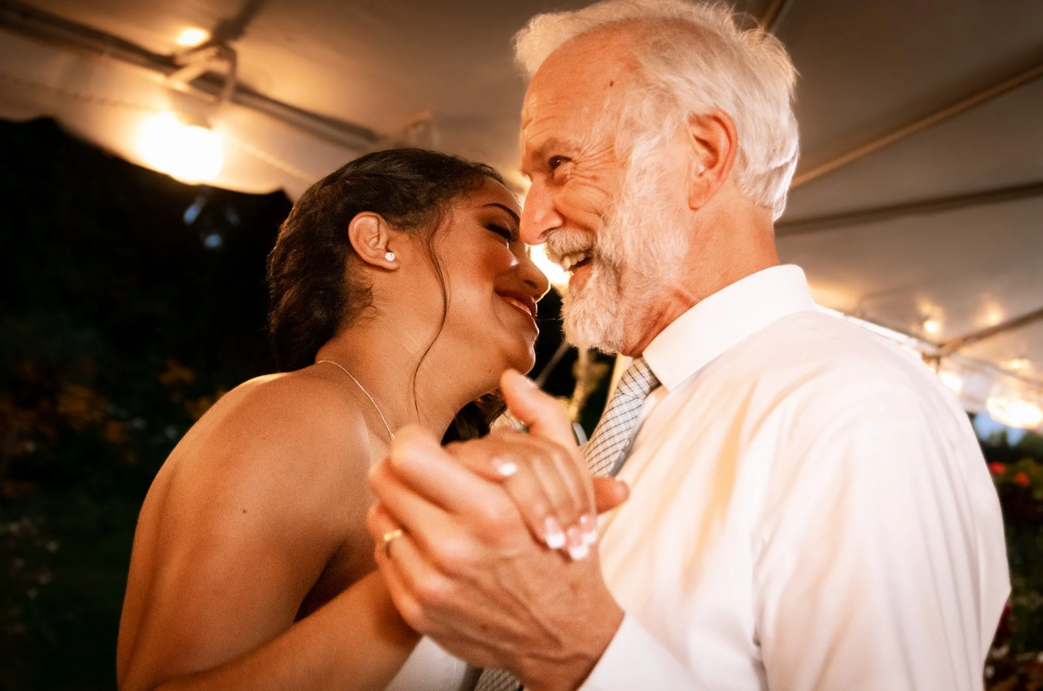 A young woman and an elderly man dancing closely, smiling and touching foreheads during a celebration or event under a tent.