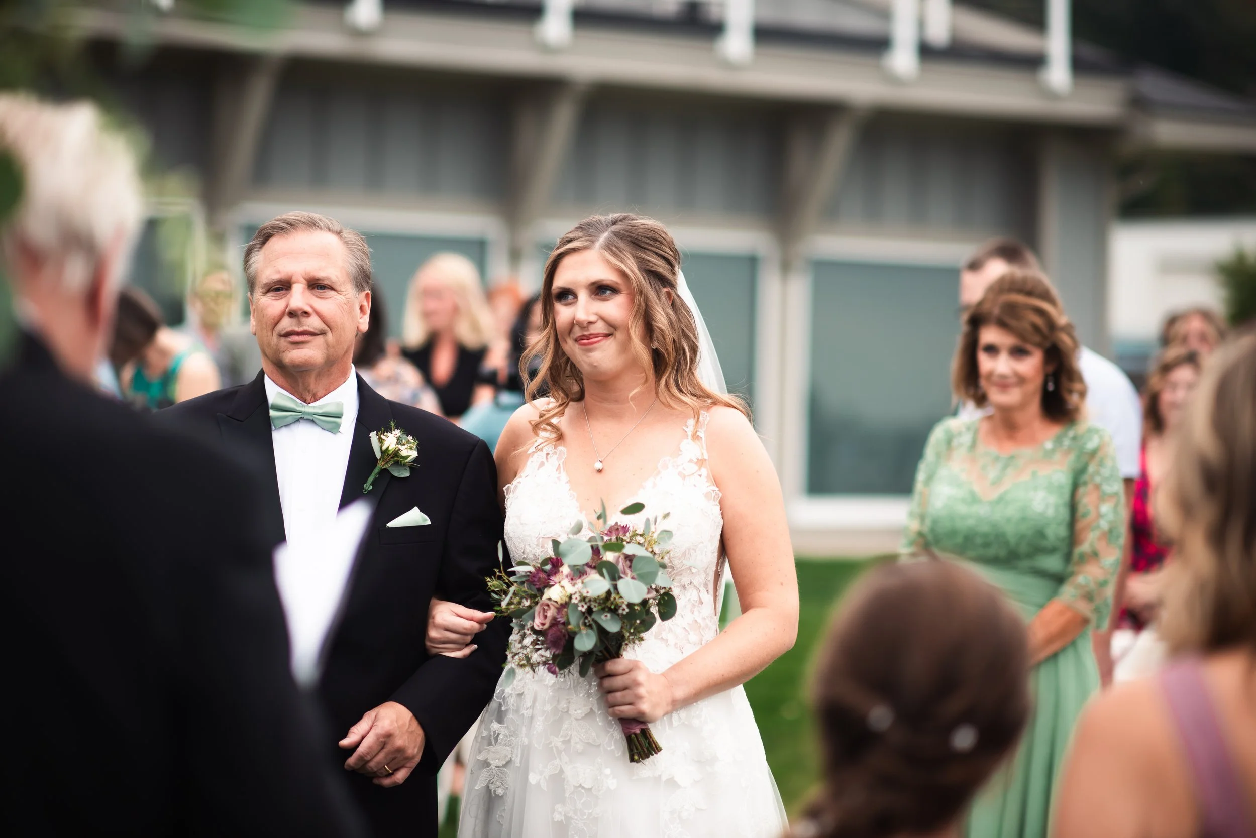 A bride in a white lace wedding dress holding a bouquet, walking down the aisle with her father during an outdoor wedding ceremony. Guests are seated and watching in the background.
