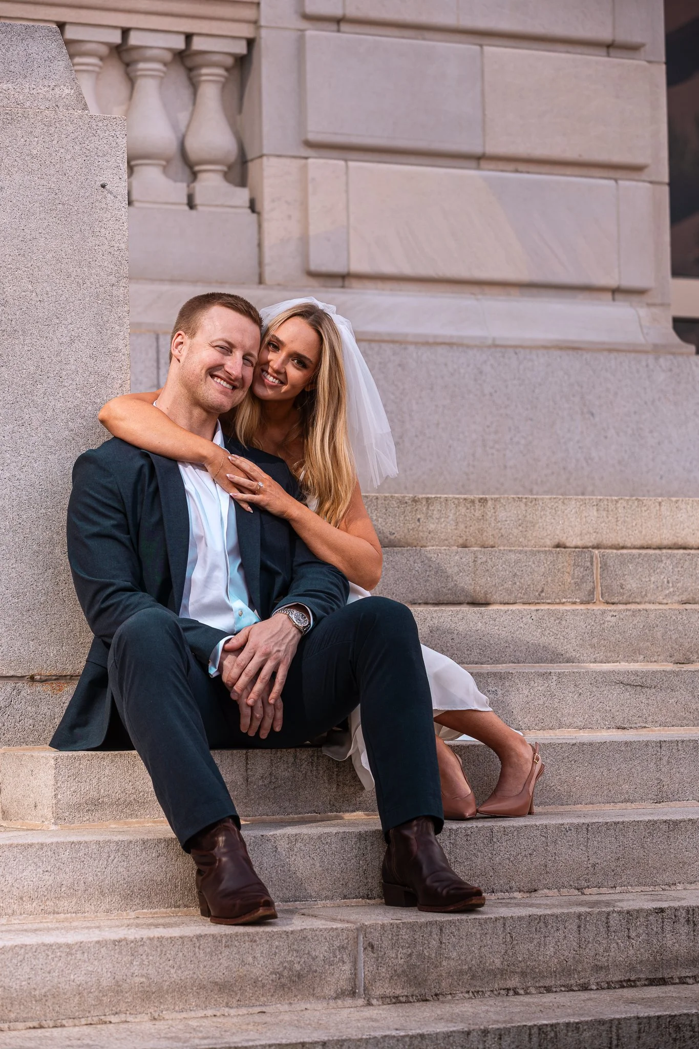 A newlywed couple sitting on stone steps in front of a historic building, smiling and embracing. The woman is wearing a wedding dress and veil, and the man is in a suit with brown boots.