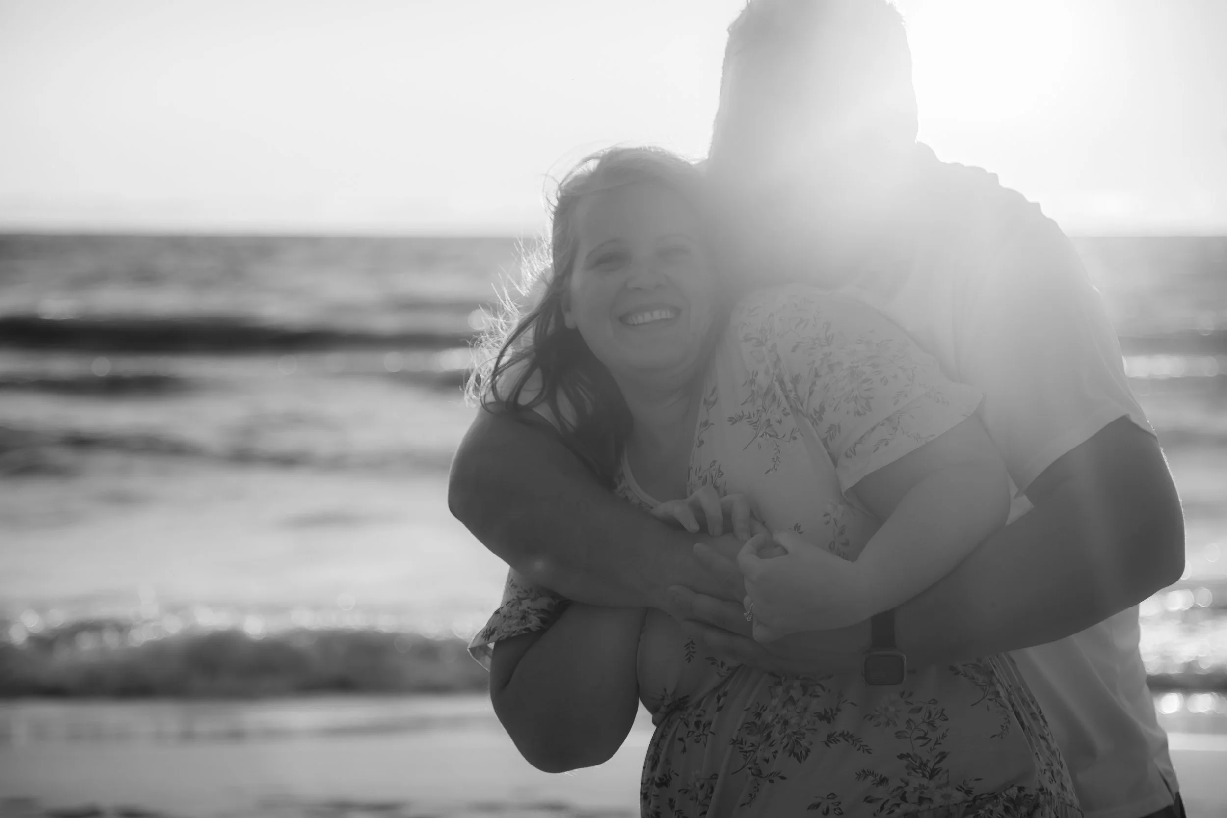 A couple hugging on the beach with the sun setting behind them, creating a backlit silhouette in black and white.