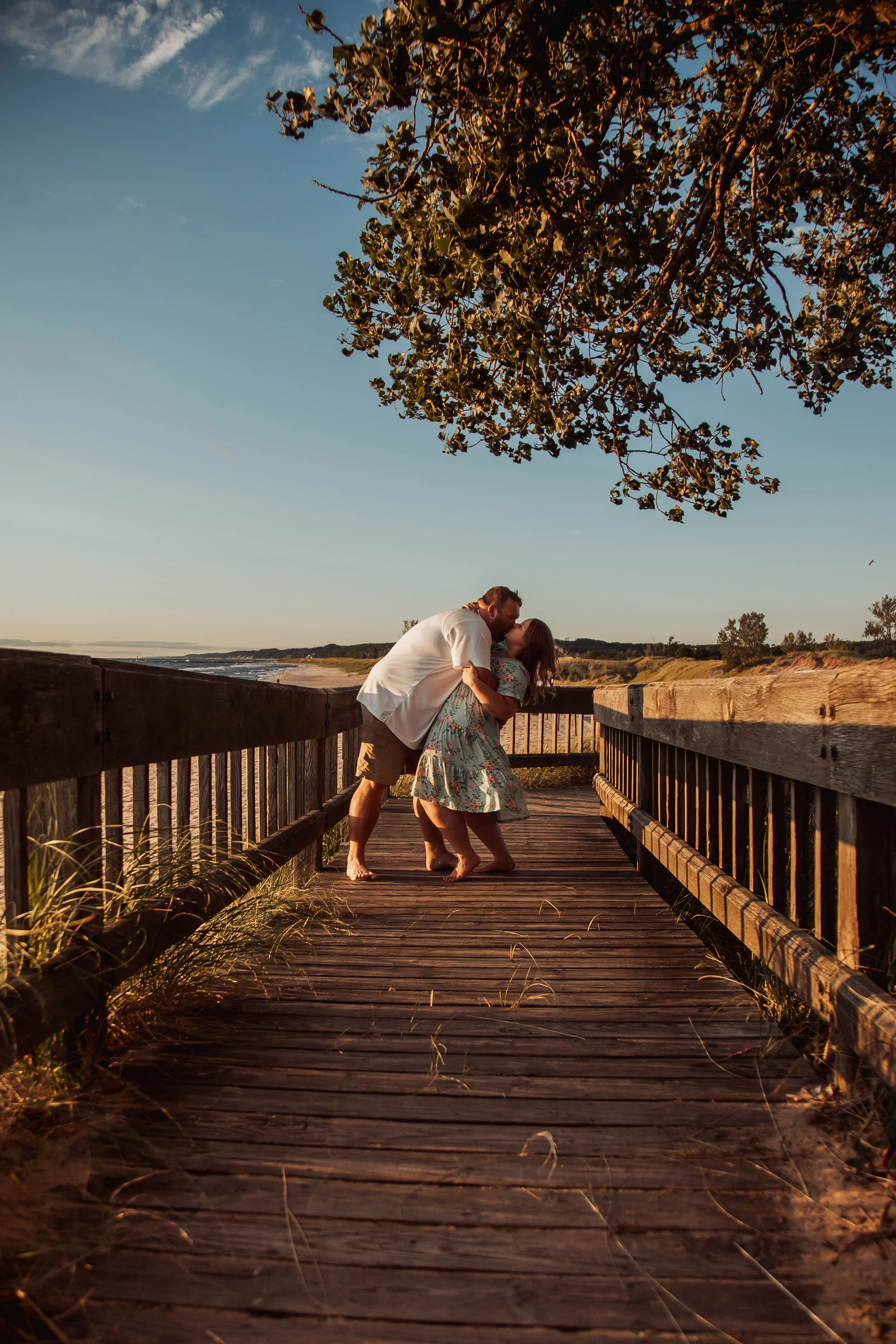 A couple kissing and embracing on a wooden bridge at sunset, with trees and a beach in the background.