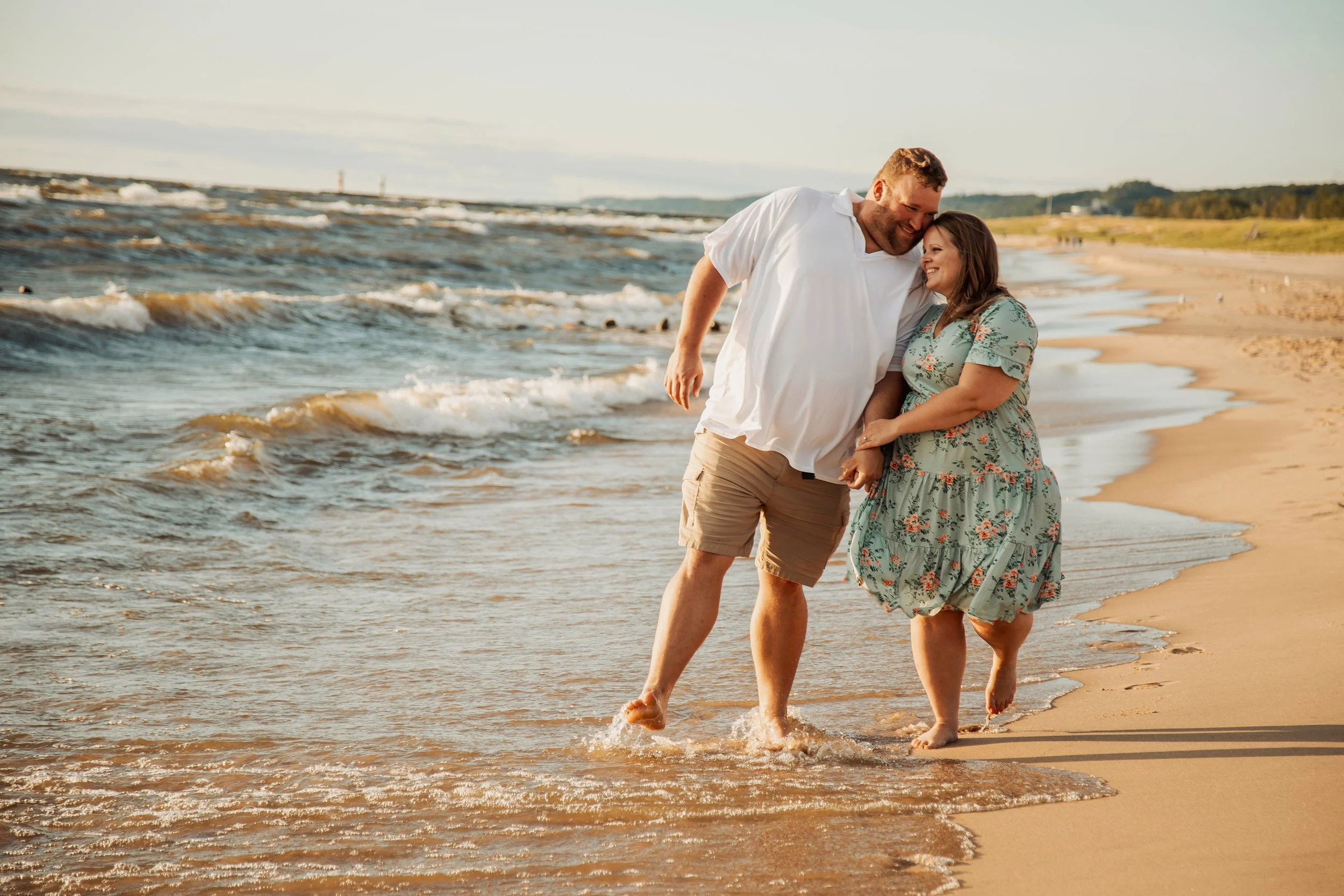 A couple holding hands and smiling at each other while walking along the beach, with gentle waves and a distant horizon.