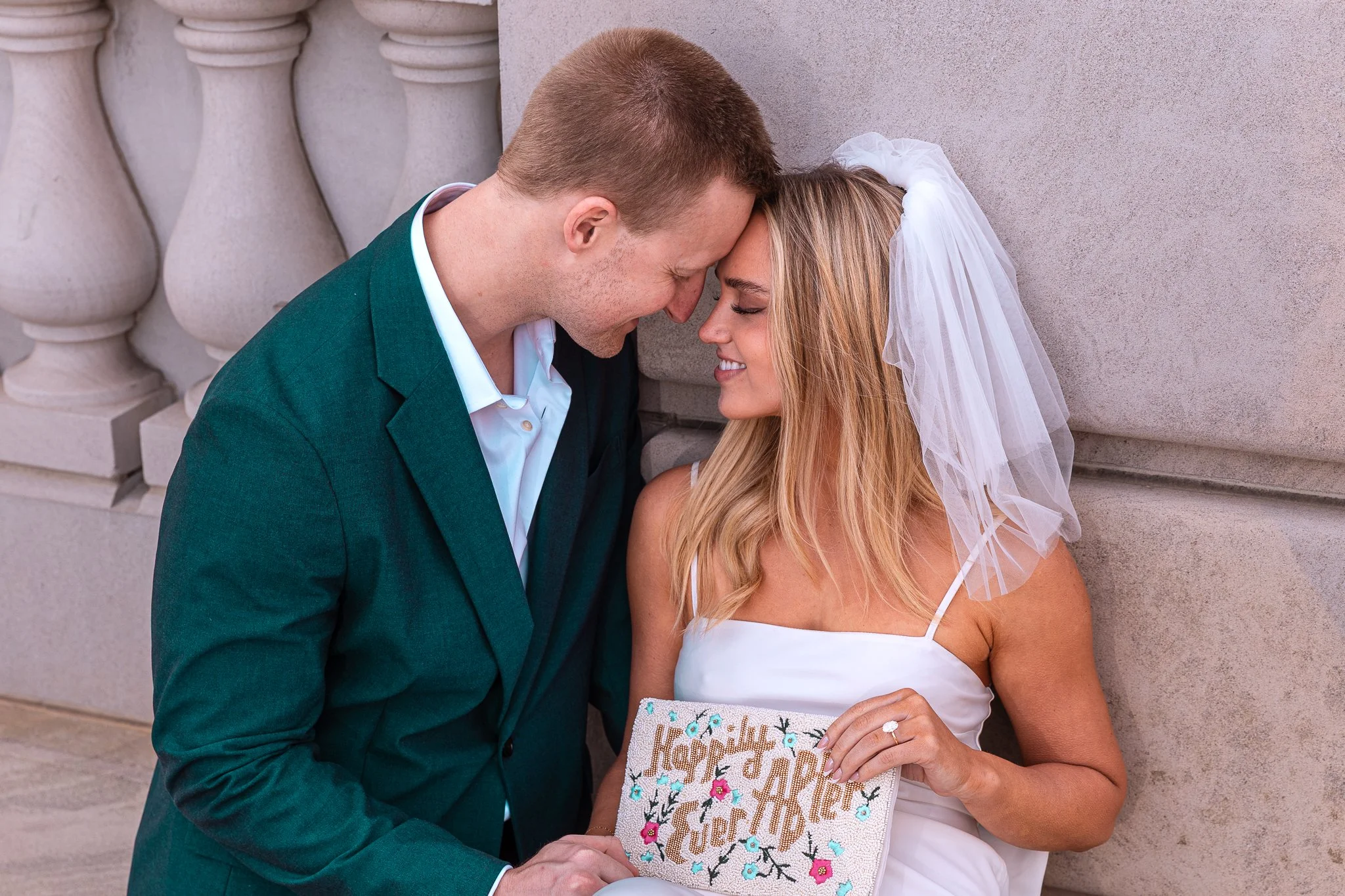 A couple dressed in wedding attire, with foreheads touching, smiling while holding a beaded card that says "Happily Ever After" in front of a stone wall with decorative balusters.