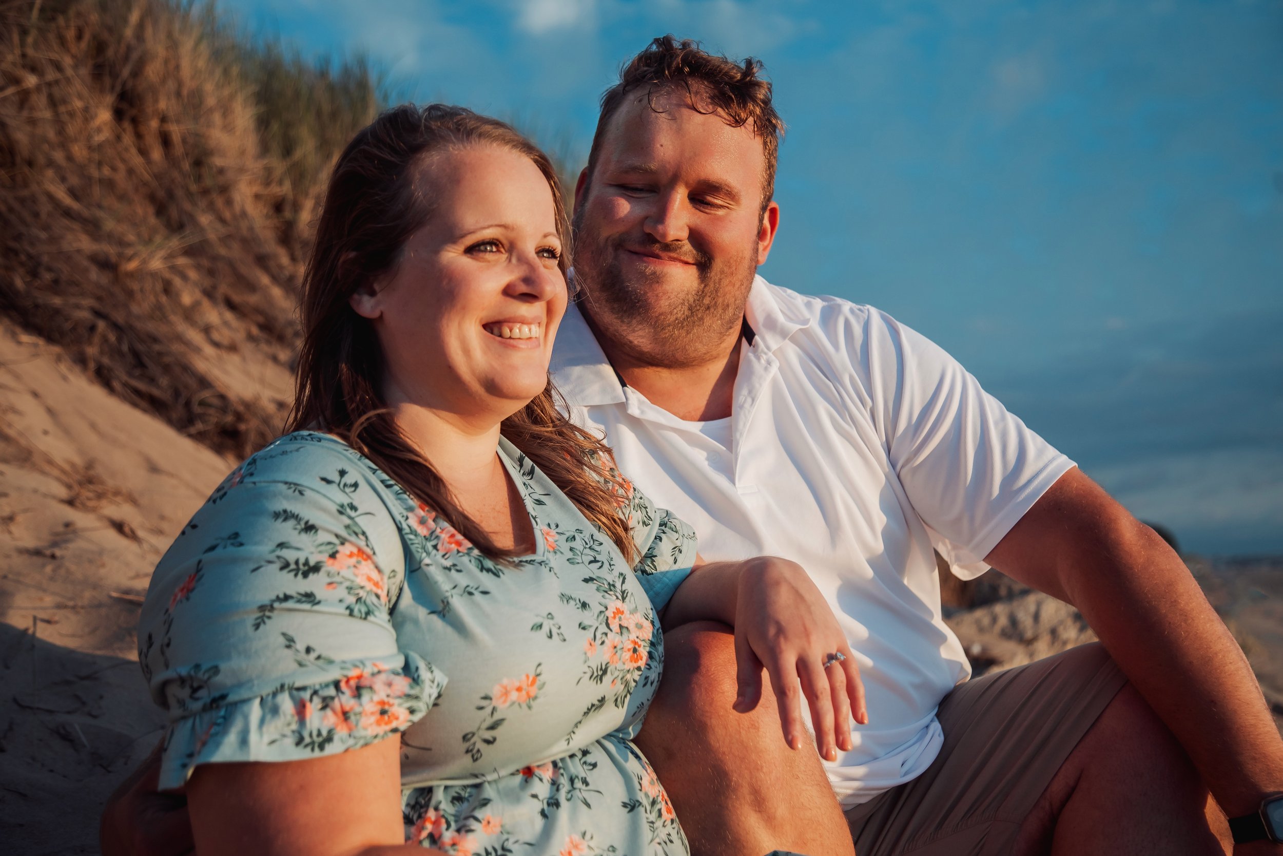 A couple sitting on a sandy beach at sunset, smiling and looking at each other.