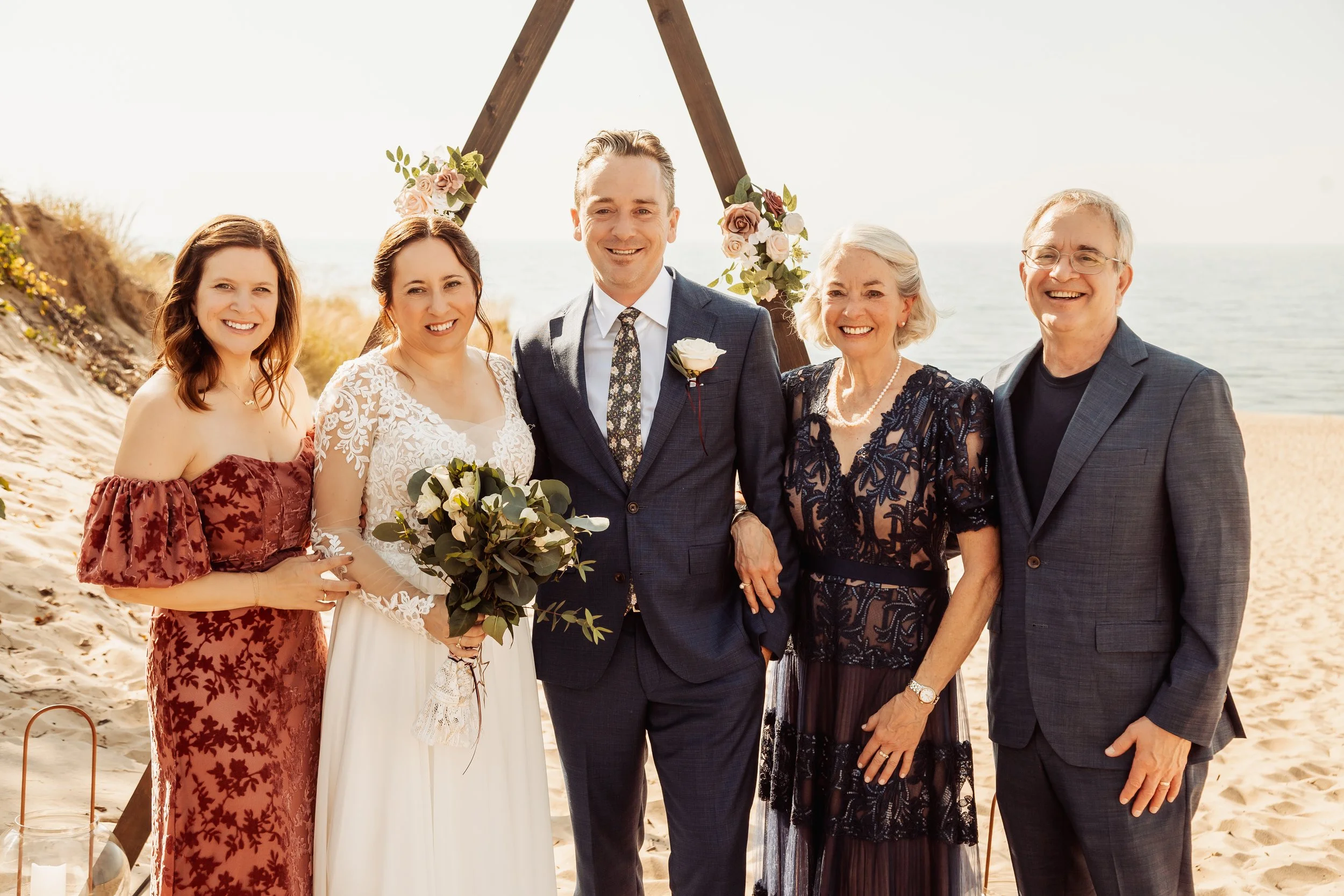 A group of five people at a beach wedding, including a bride holding a bouquet, a groom, and three other family members or friends standing beside them in front of a wooden wedding arch decorated with flowers.