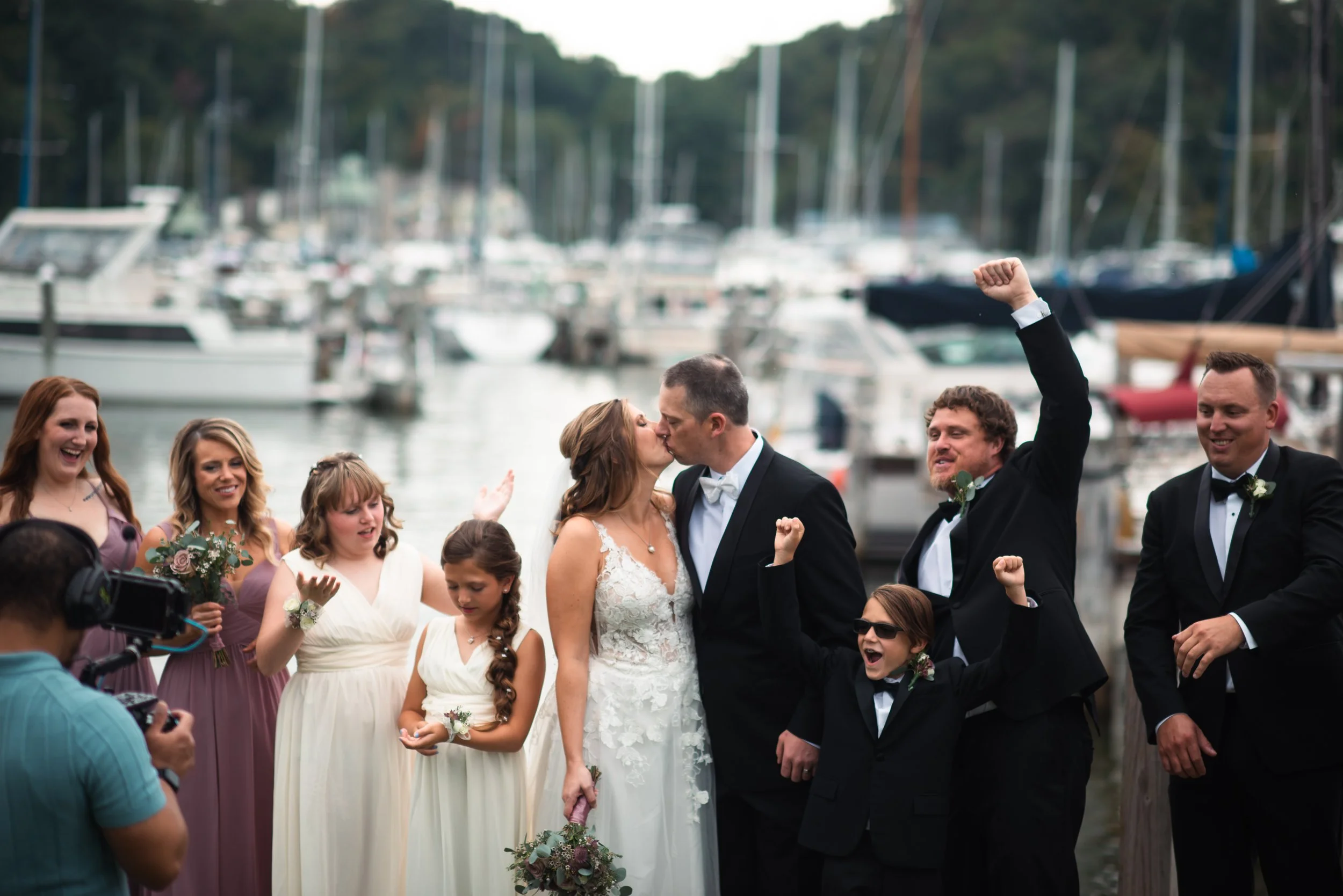 A wedding group celebration at a marina, with the bride and groom kissing and surrounded by friends and family, some cheering and one with raised fist, in front of boats.