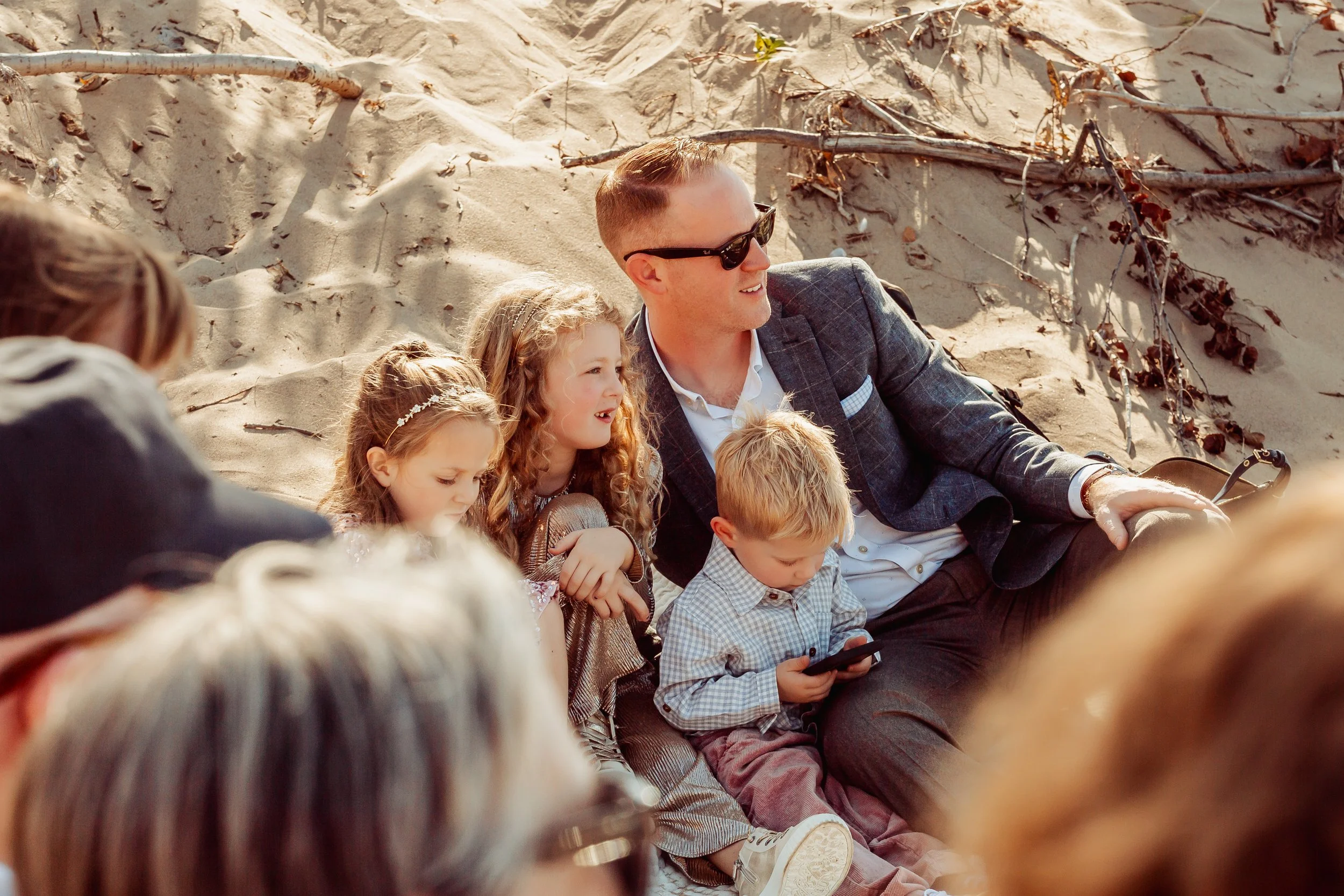 A man and four children sitting on a sandy beach, with two adults partially visible in the foreground. The man is wearing sunglasses and a blazer, and the children are engaged with a mobile phone and looking around, enjoying the outdoor setting.