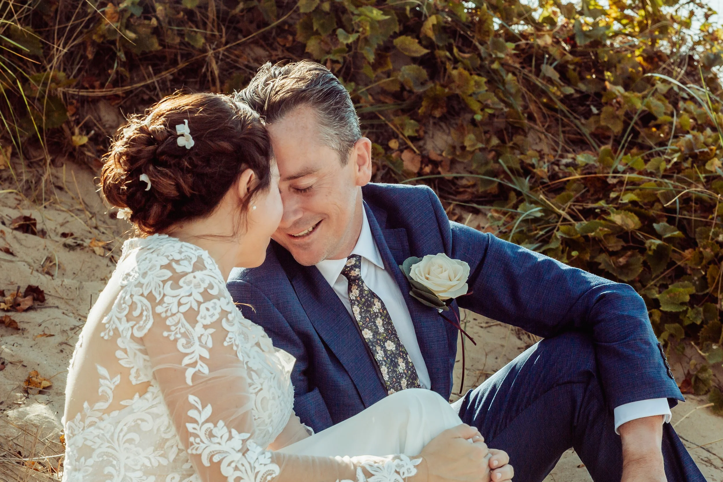 A bride and groom sitting on the sand, sharing an intimate moment with foreheads touching, surrounded by dunes and foliage.