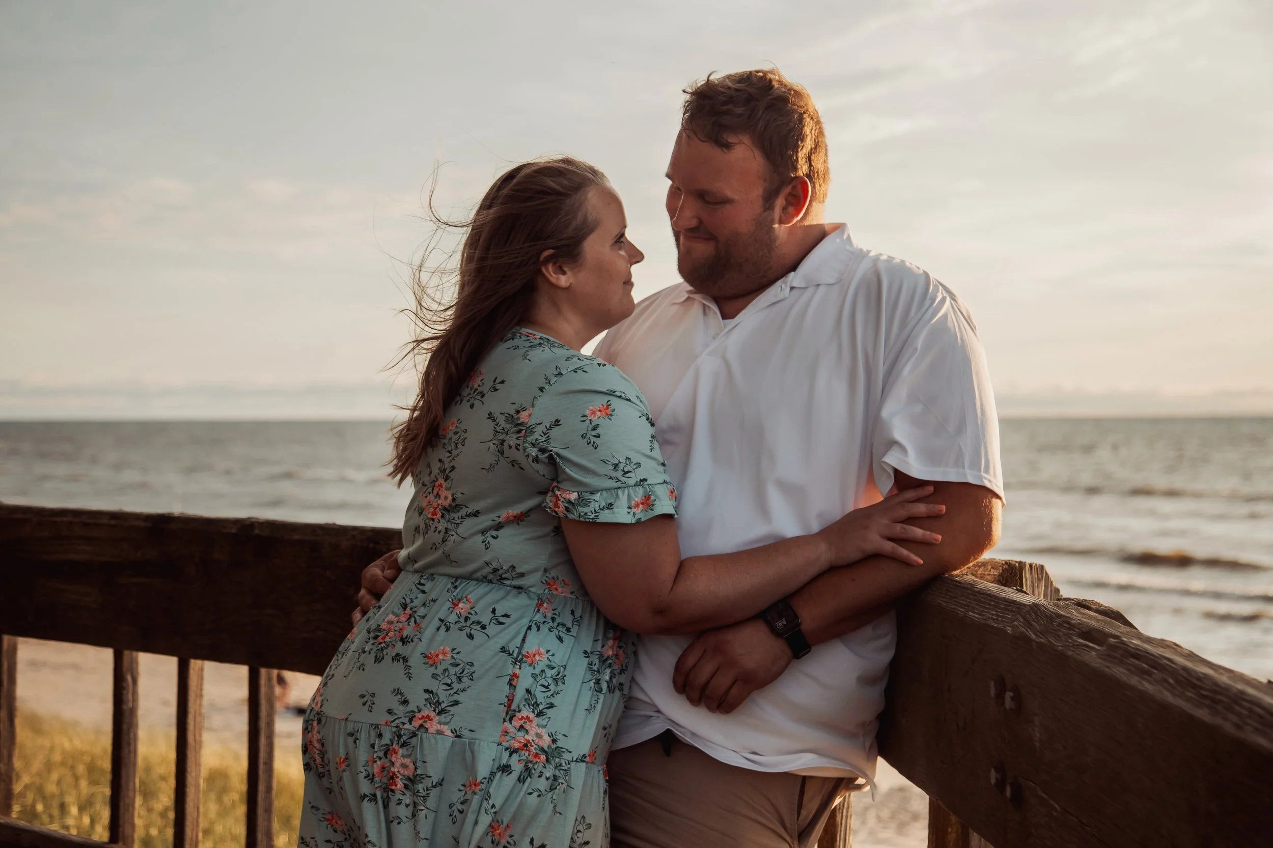 A couple standing close together on a beach at sunset, looking into each other's eyes, with the ocean and sky in the background.