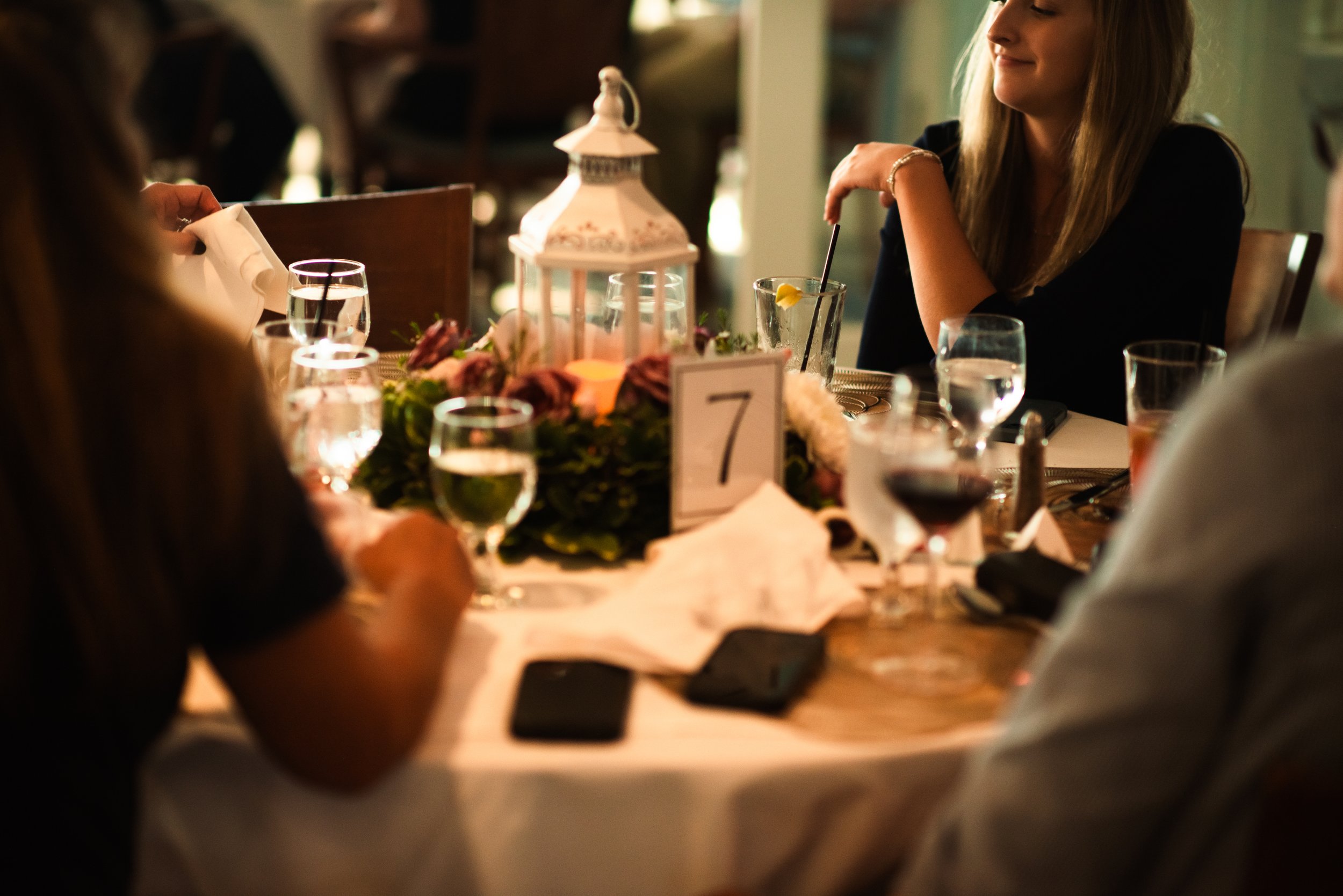 People sitting around a decorated table at a formal event, with a white lantern centerpiece, floral arrangement, table number 7, and glasses of water and wine.