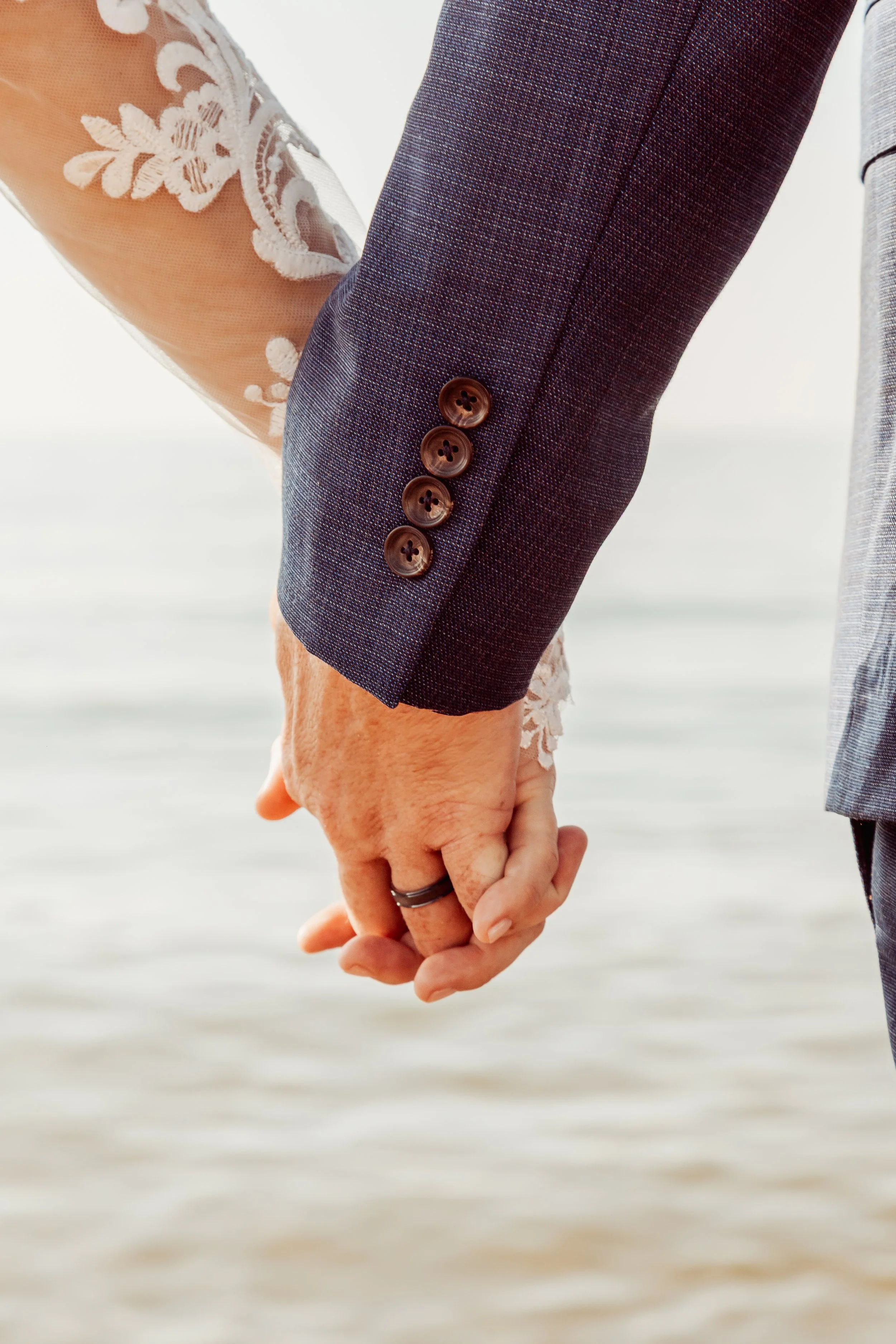 Close-up of a couple holding hands, with the man wearing a navy suit and the woman wearing a white lace dress, at the beach.