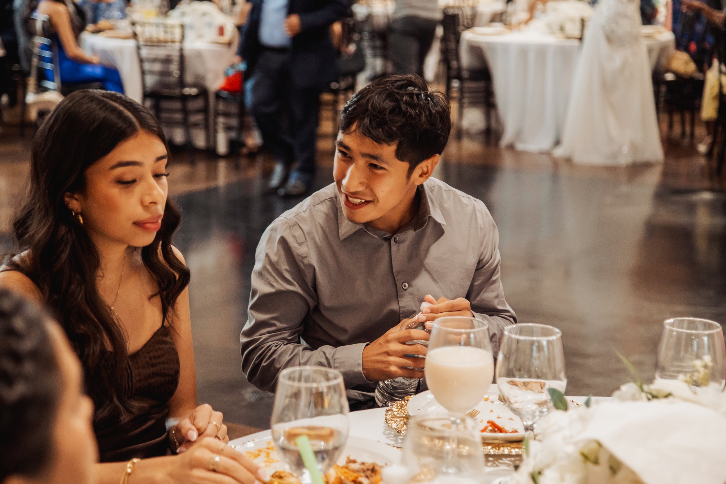 A young man and woman seated at a table during a formal dinner event, with other guests and round banquet tables in the background.