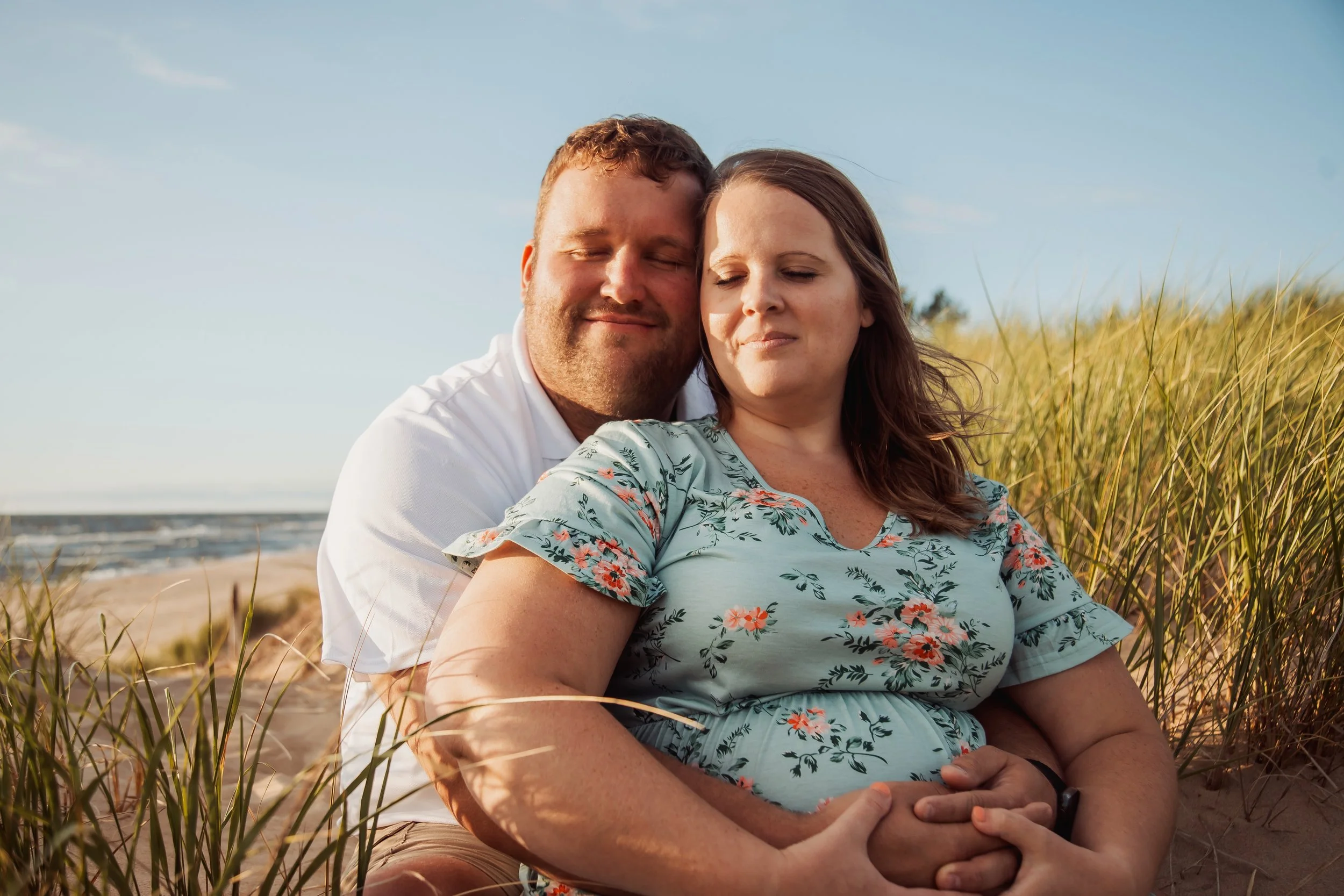 A pregnant woman and a man sitting on the beach, hugging, with tall grass, sandy beach, and ocean in the background, during daylight.