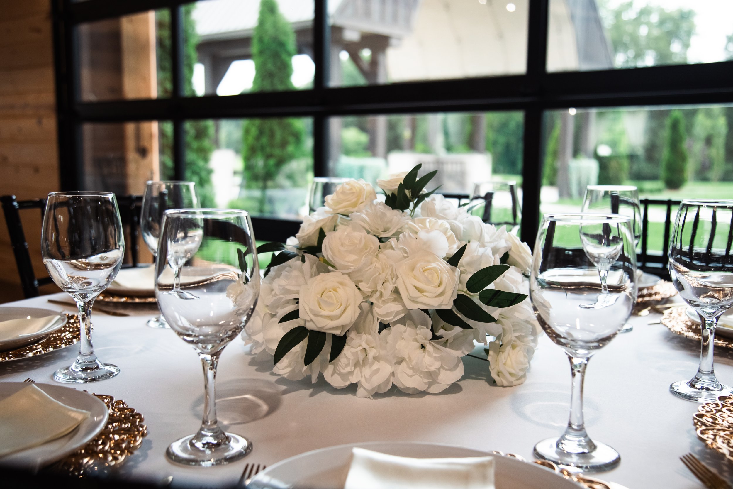 Elegant dining table with a large white floral centerpiece, wine glasses, and place settings, set inside a rustic room with large windows overlooking a green outdoor area.