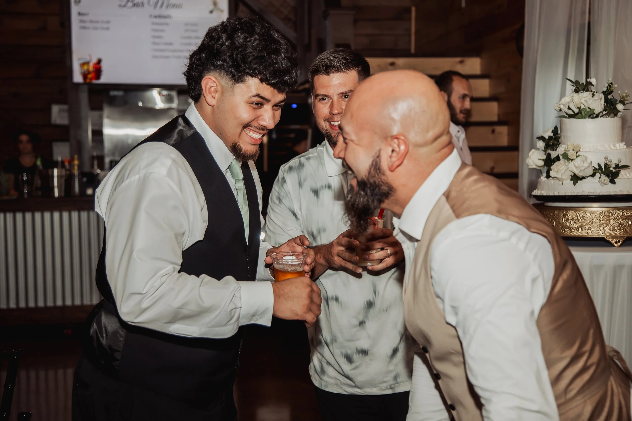 Four men laughing and talking at a social event, with drinks in hand, in a rustic wood-paneled room with a large wedding cake decorated with white flowers in the background.