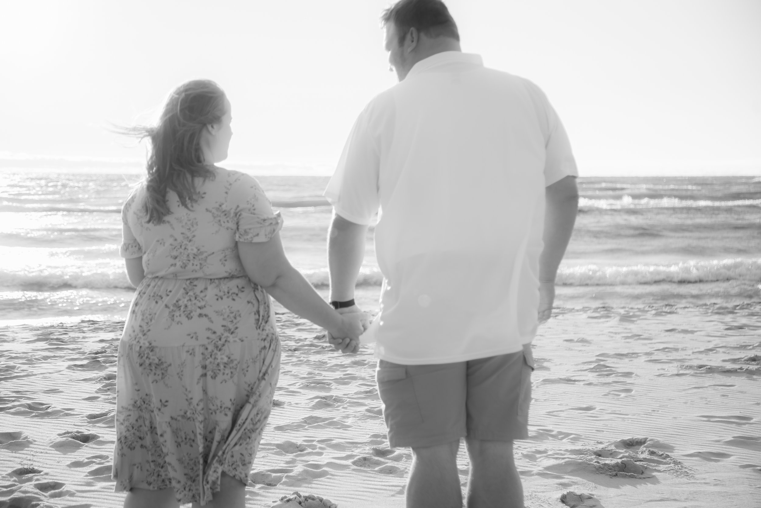 A couple holding hands walking on the beach, facing the ocean at sunset in black and white.