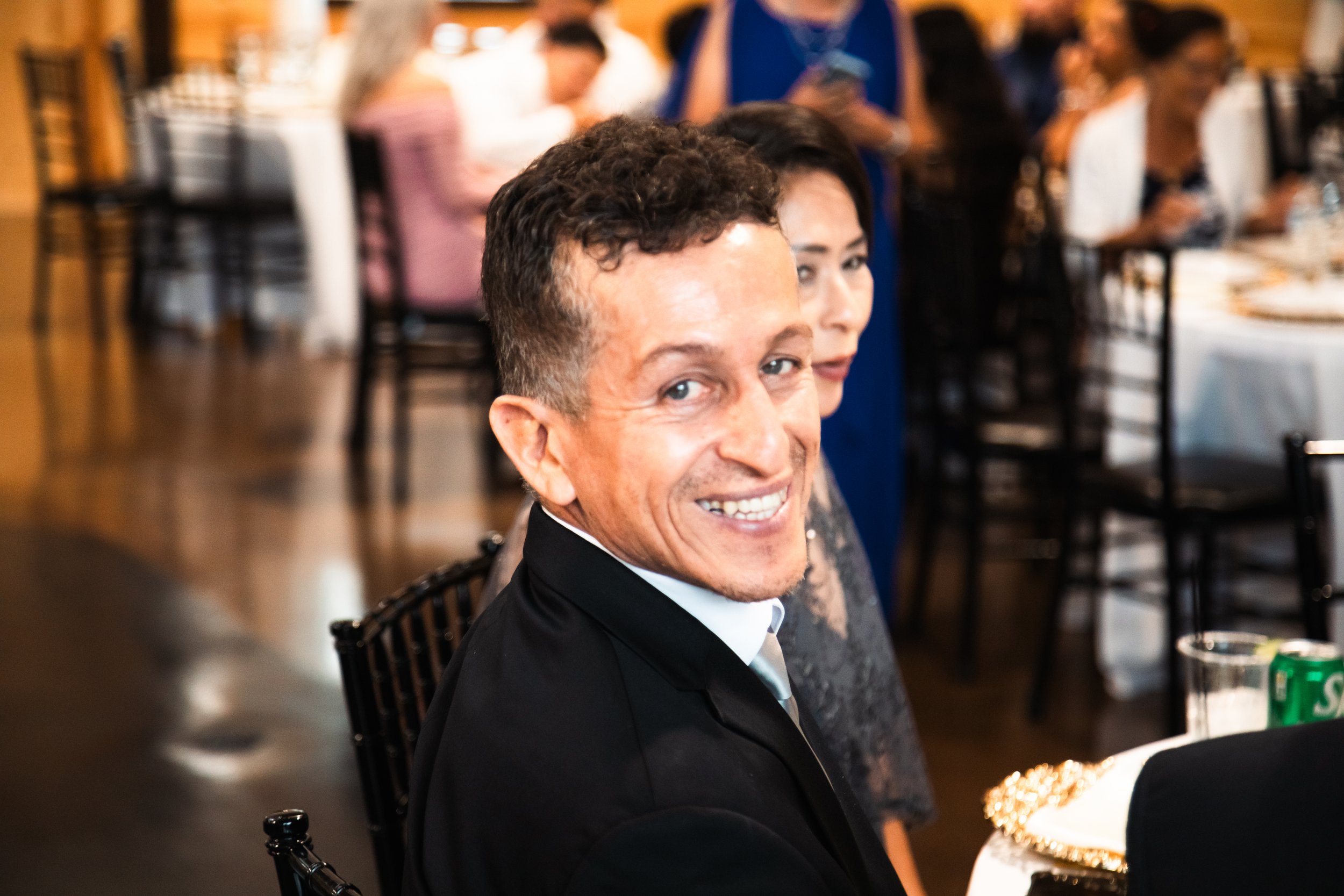 A man with short, curly dark hair smiling at a formal event. He is dressed in a black tuxedo with a white shirt and a grey tie. A woman with dark hair and a patterned dress sits beside him. The background shows other guests seated at tables with whit