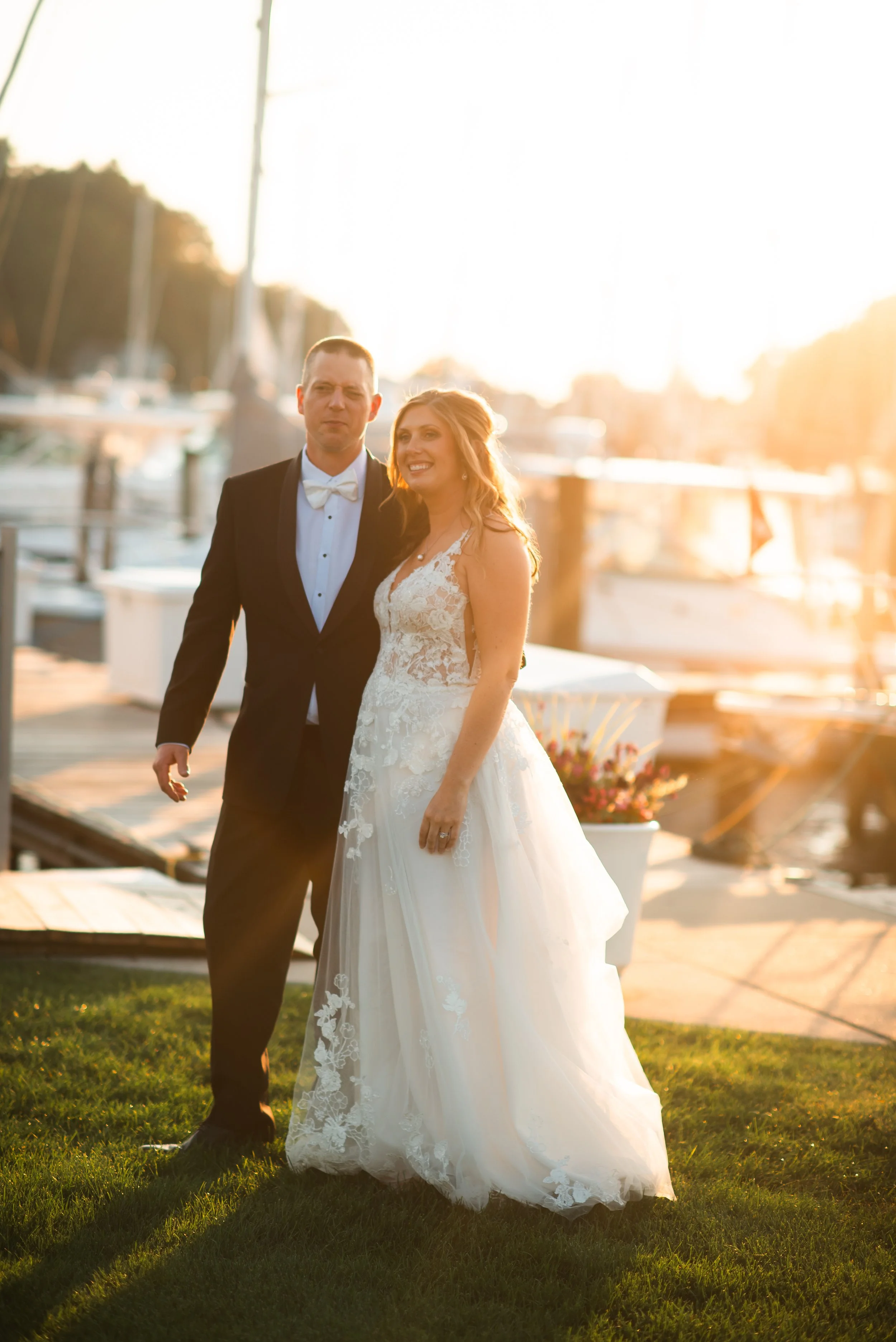 A newlywed couple standing on grass near a marina at sunset, with boats and water in the background. The groom is in a black tuxedo with a white bow tie, and the bride is in a lace wedding dress. They are smiling and posing for a photo.