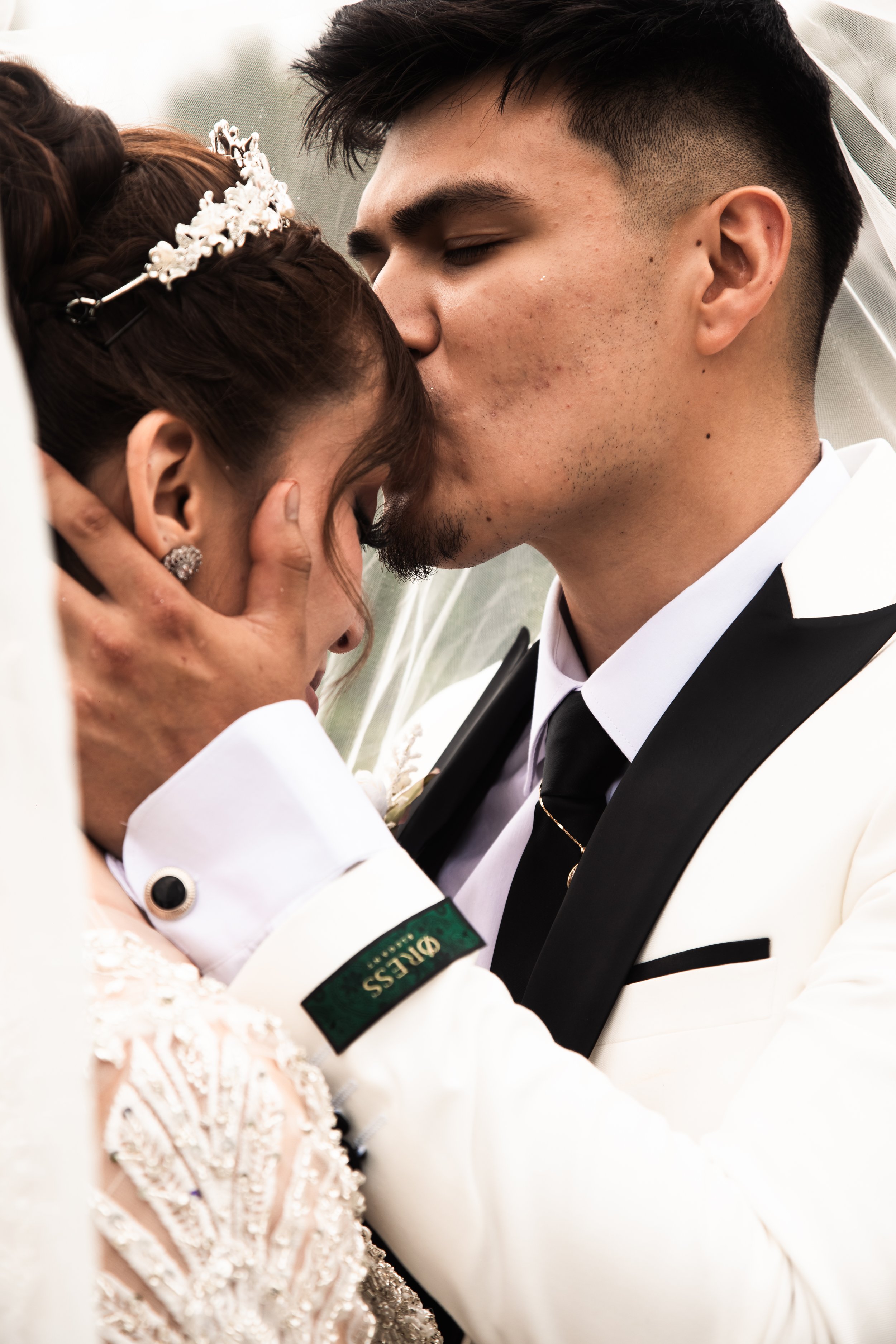 A man and woman in wedding attire share a kiss, with the man gently holding the woman's face and both in formal wedding clothing.