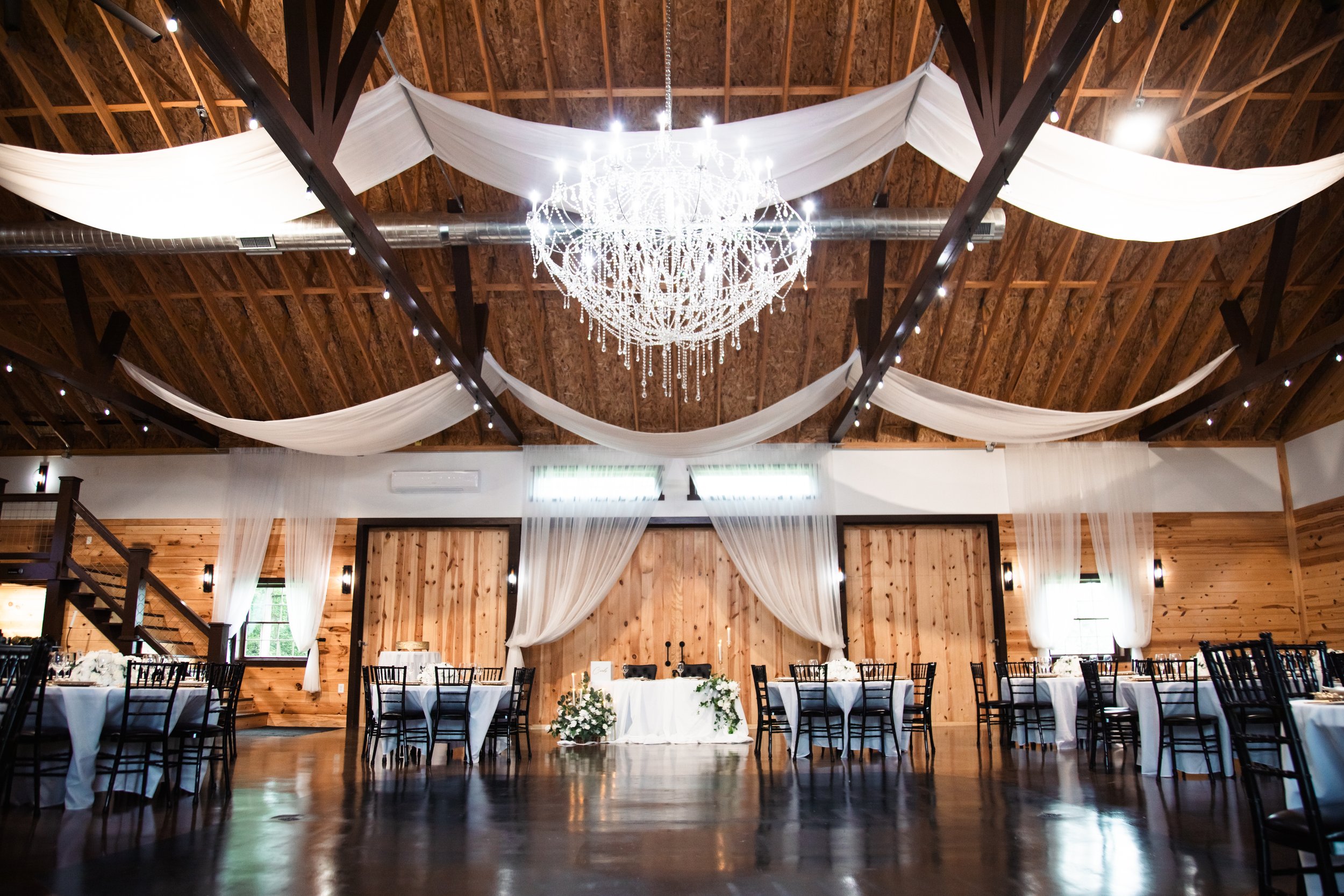 A decorated event hall with a high wooden ceiling, white draped fabric, a large chandelier, and round tables set for a formal occasion.