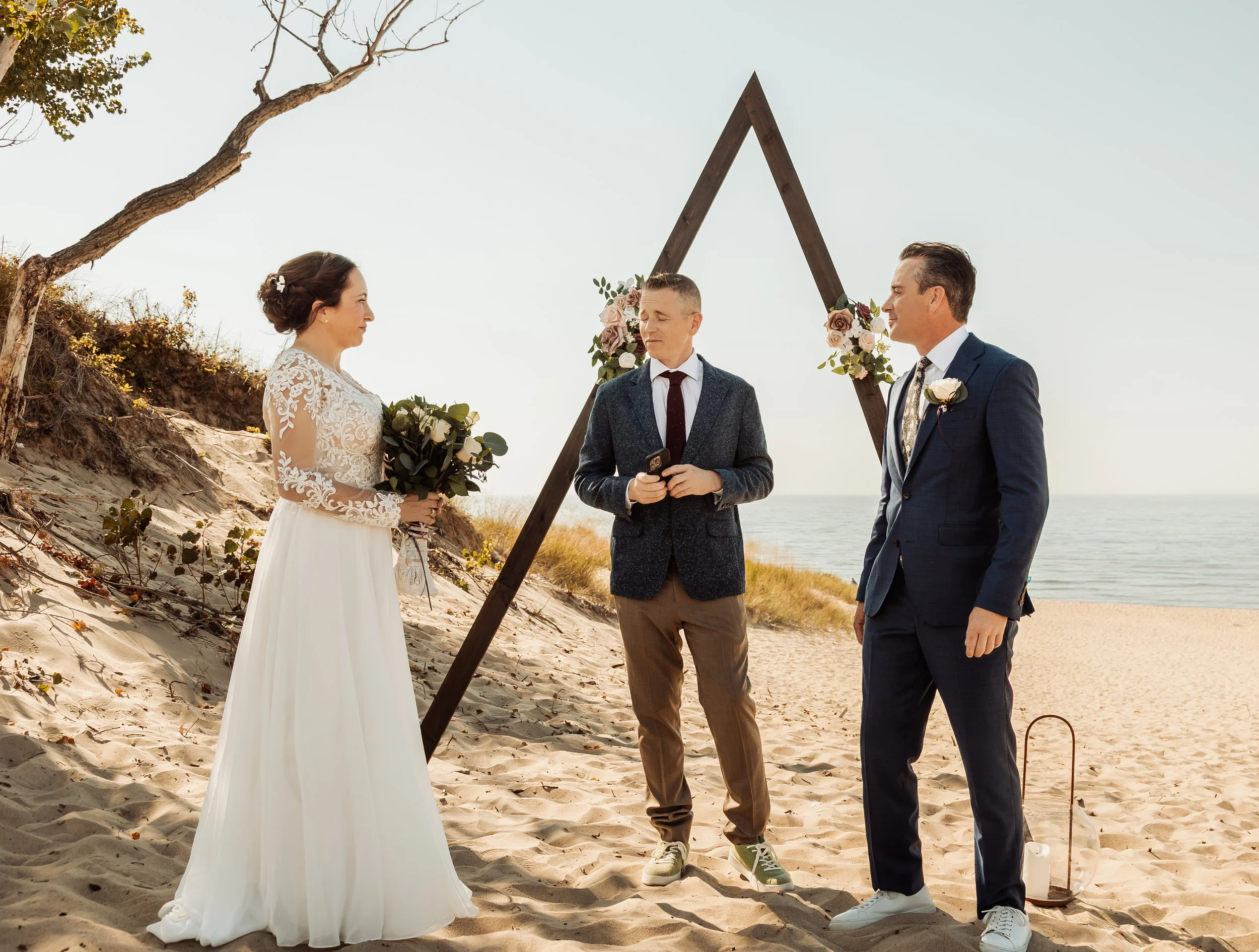 A couple getting married on a beach, standing under a wooden triangular wedding arch decorated with pink and white flowers. The bride is holding a bouquet of flowers, and the groom is wearing a suit with a boutonniere. An officiant, holding a phone, 