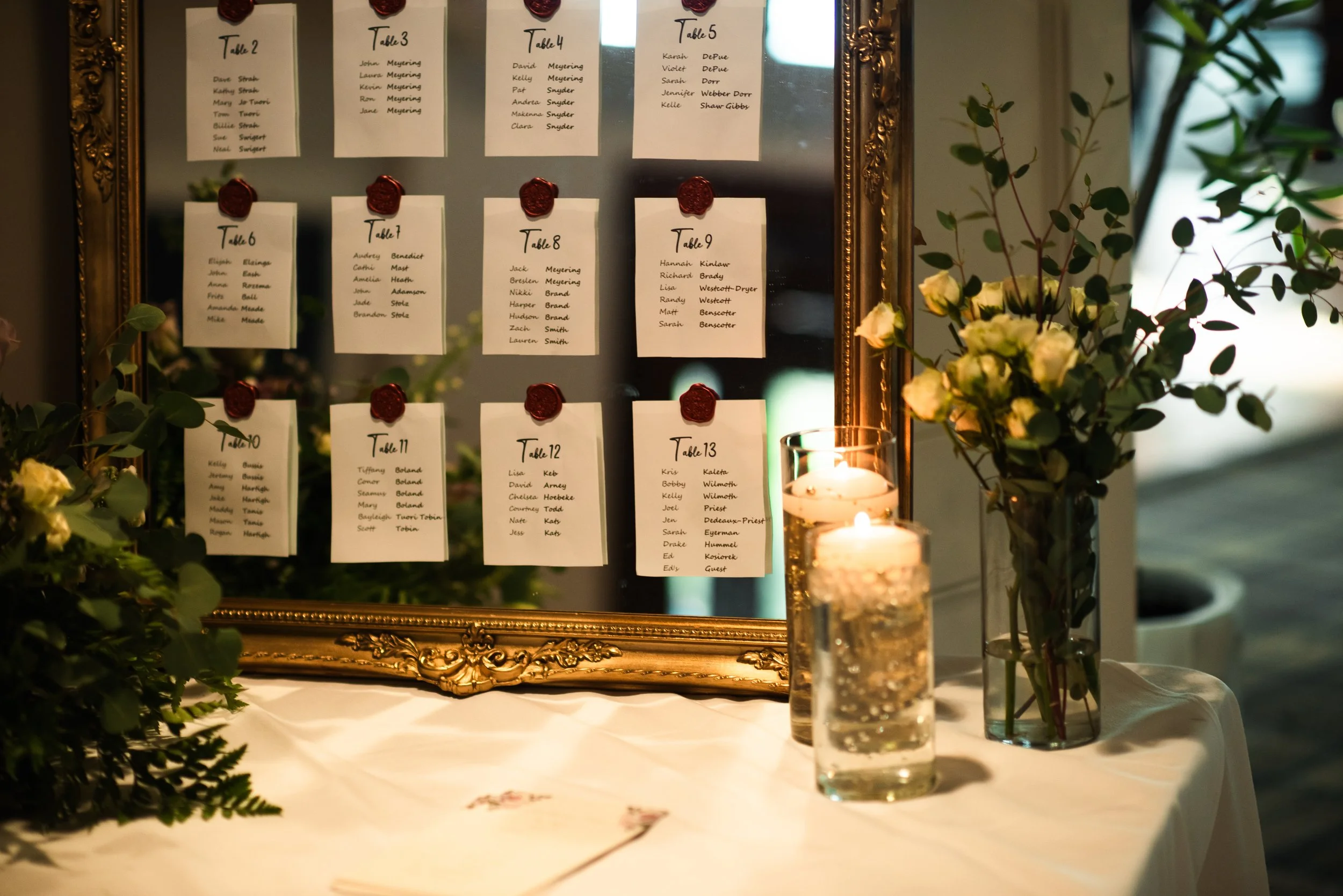 A wedding seating chart with white cards pinned on an ornate gold-framed mirror, surrounded by lit candles and flower arrangements, displaying table assignments and guest names.