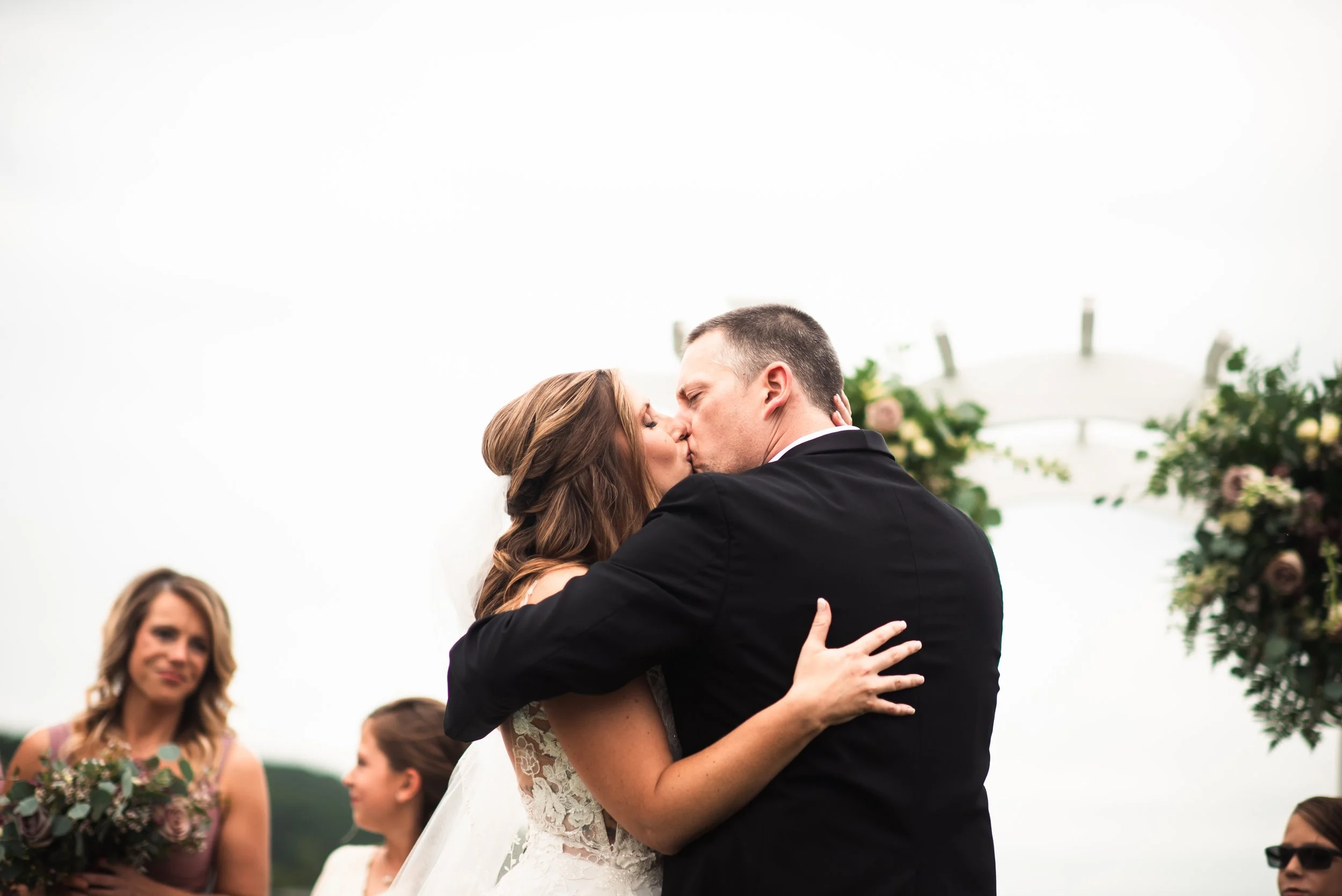 A couple is kissing at their wedding ceremony outdoors, with friends and a decorated arch in the background.