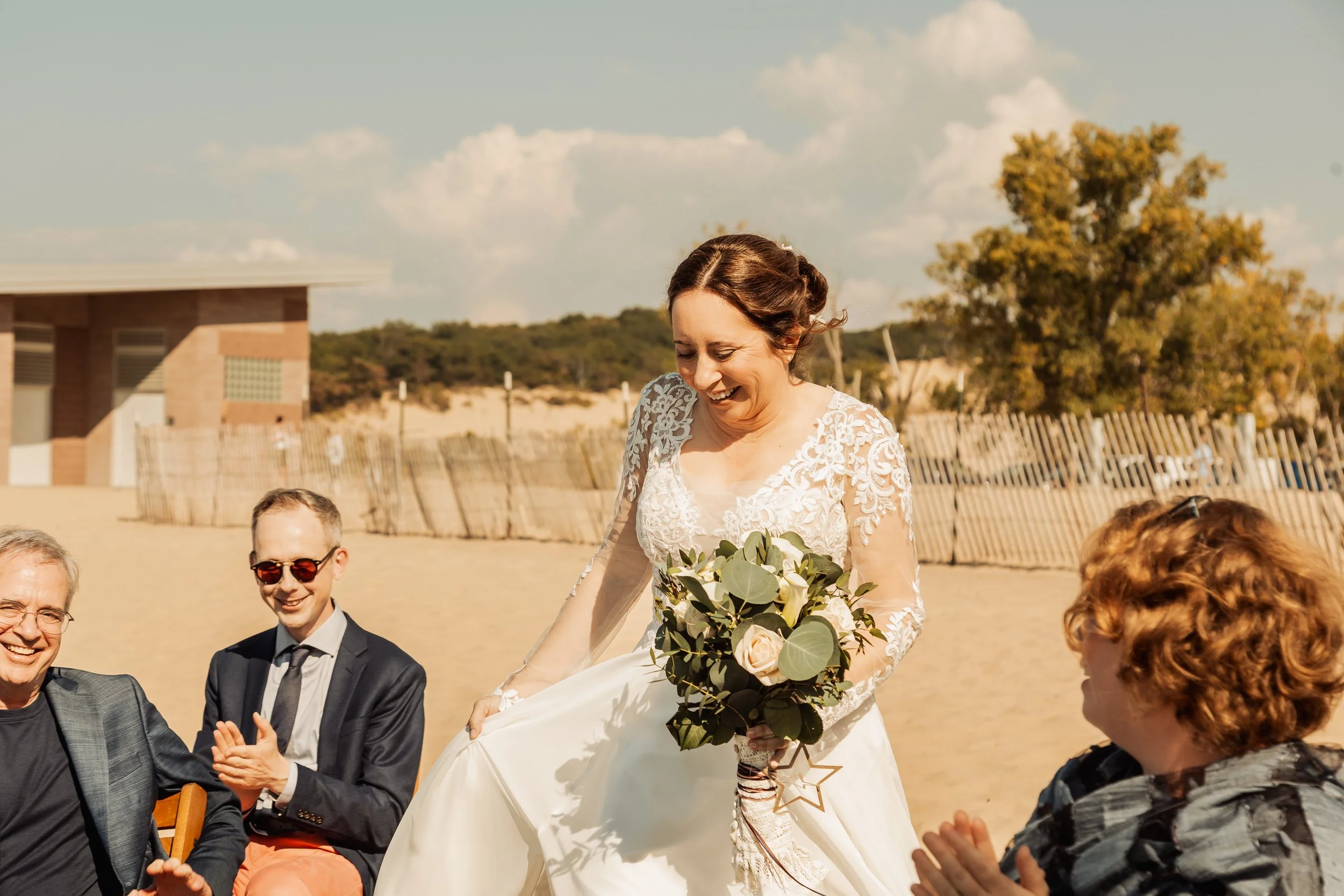 A bride in a white lace wedding dress holding a bouquet smiling down at seated guests on a sandy beach, with a backdrop of a wooden fence, trees, and a building under a partly cloudy sky.