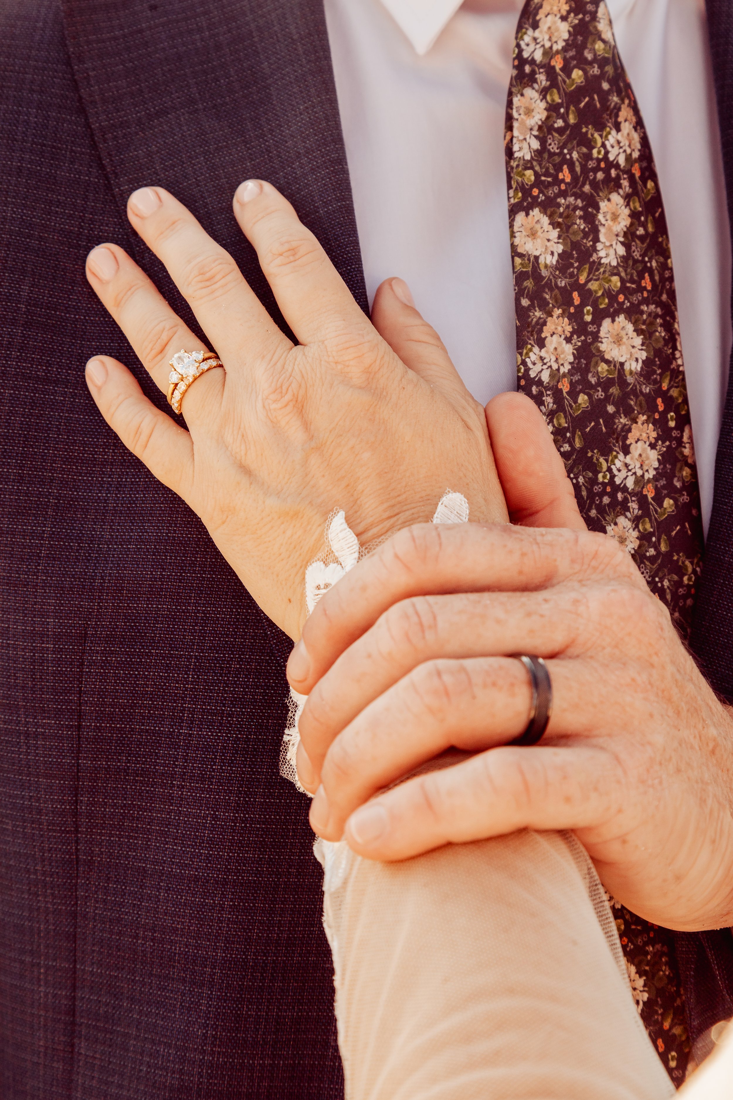 Close-up of a woman and man holding hands. The woman's hand, wearing a diamond ring, is resting on the man's chest, who is wearing a wedding band. The man is in a suit with a floral tie, and the woman has a lace sleeve.