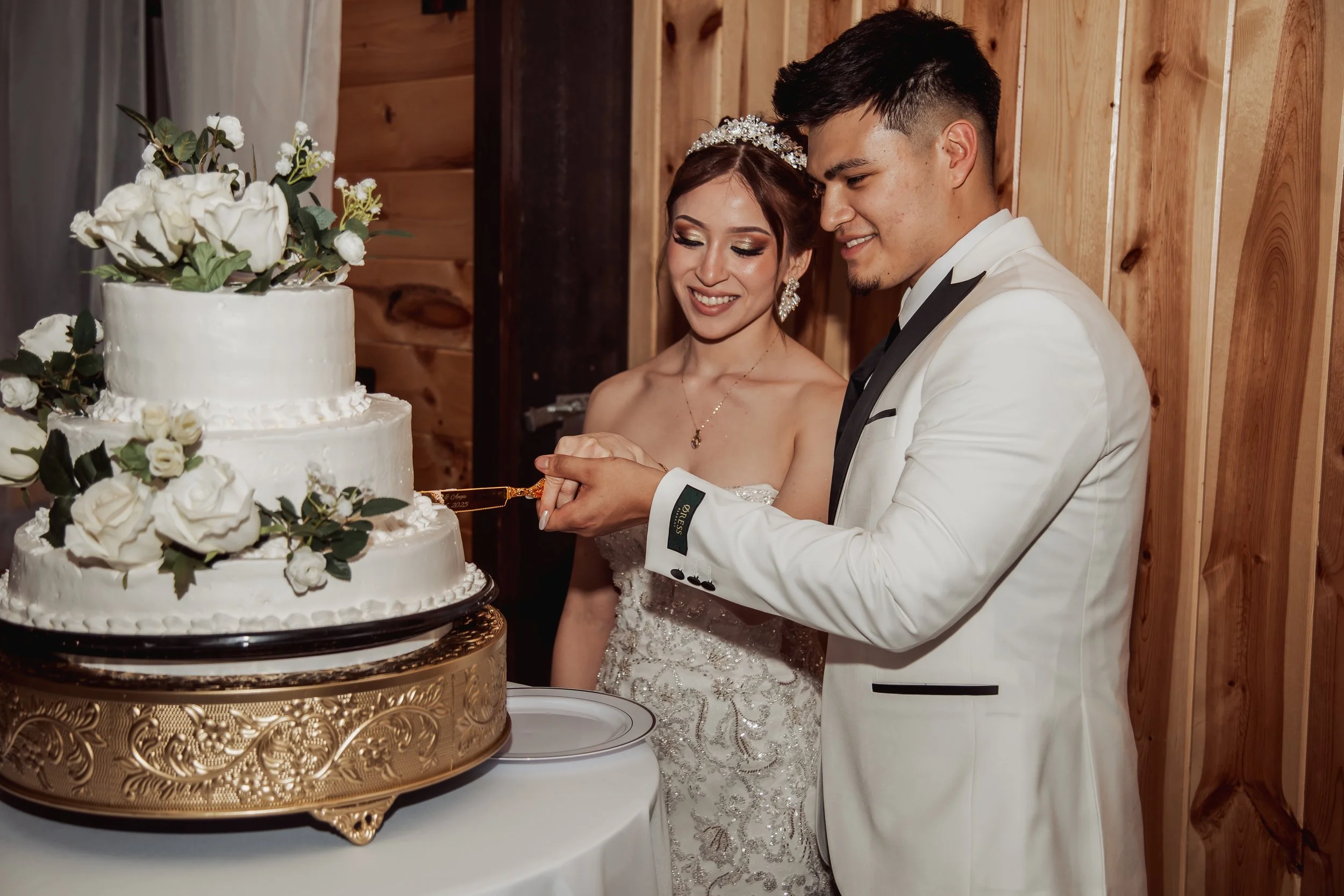 Newlywed couple cutting a wedding cake together, with the bride smiling and the groom holding a cake knife, in front of a three-tiered white wedding cake decorated with white flowers and greenery, set on a decorative gold cake stand.