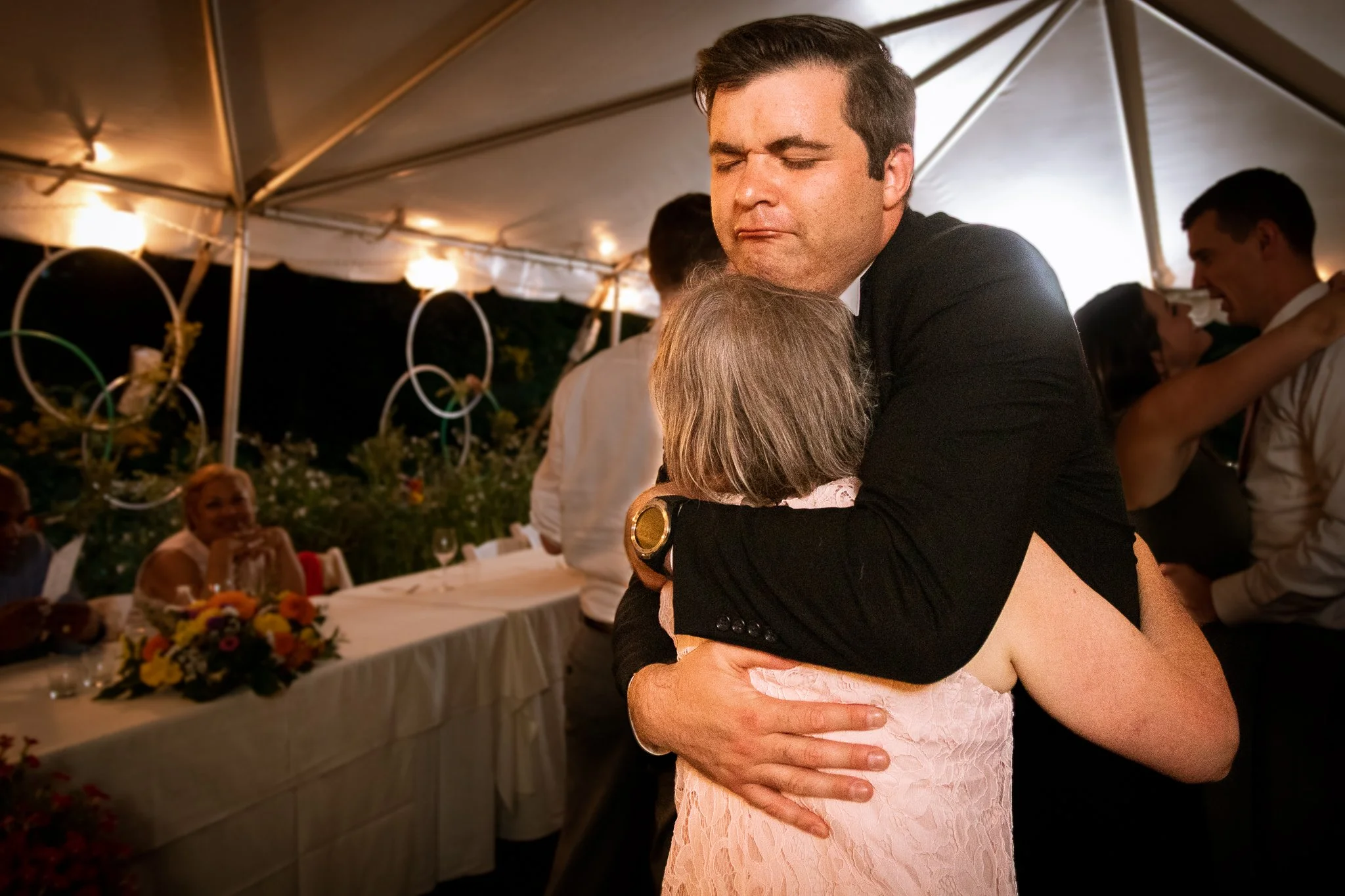 A man and a woman hugging each other at a celebration or party under a tent, with other people seated at tables in the background.