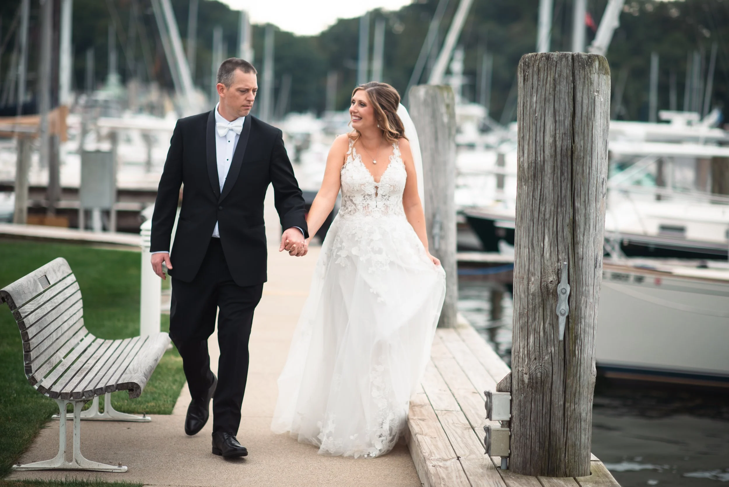 Bride and groom walking hand-in-hand along a dock, with boats in the background.