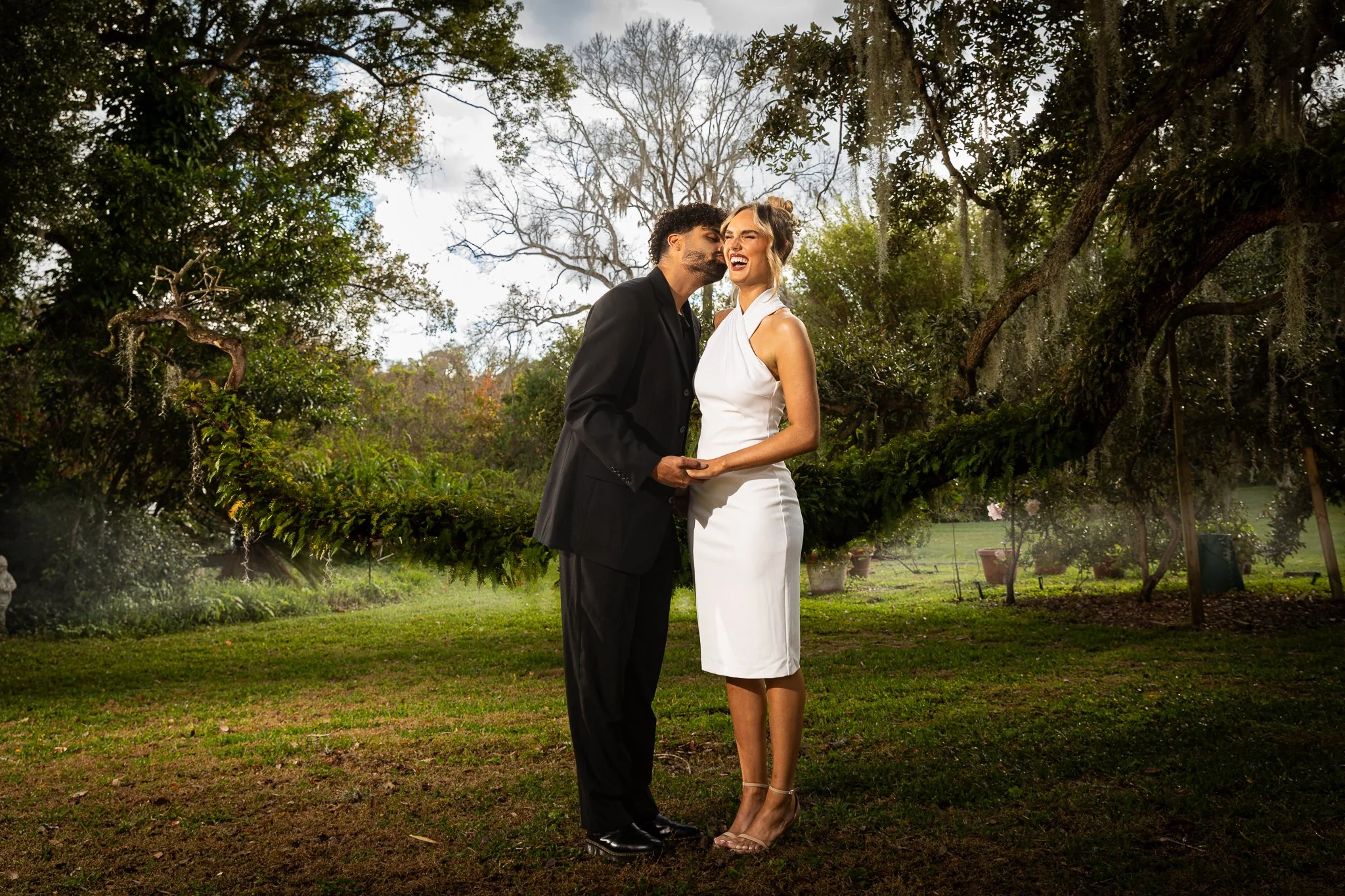 A happy couple in wedding attire holding hands, standing outdoors under a large, moss-draped tree, in a lush garden setting.
